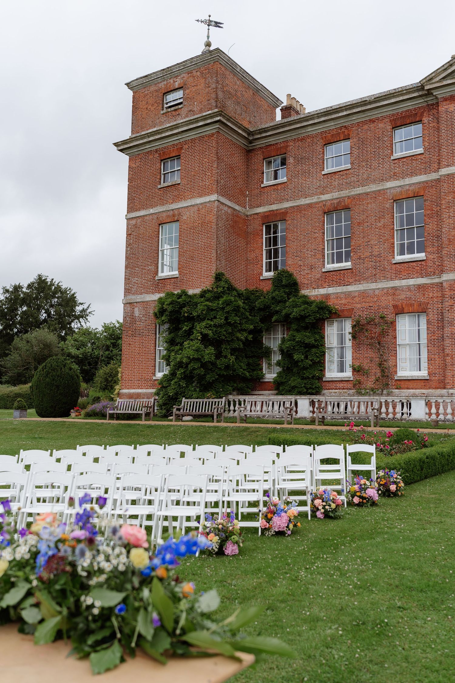 White wedding chairs arranged on lawn before grand red brick English country house with colorful flowers.