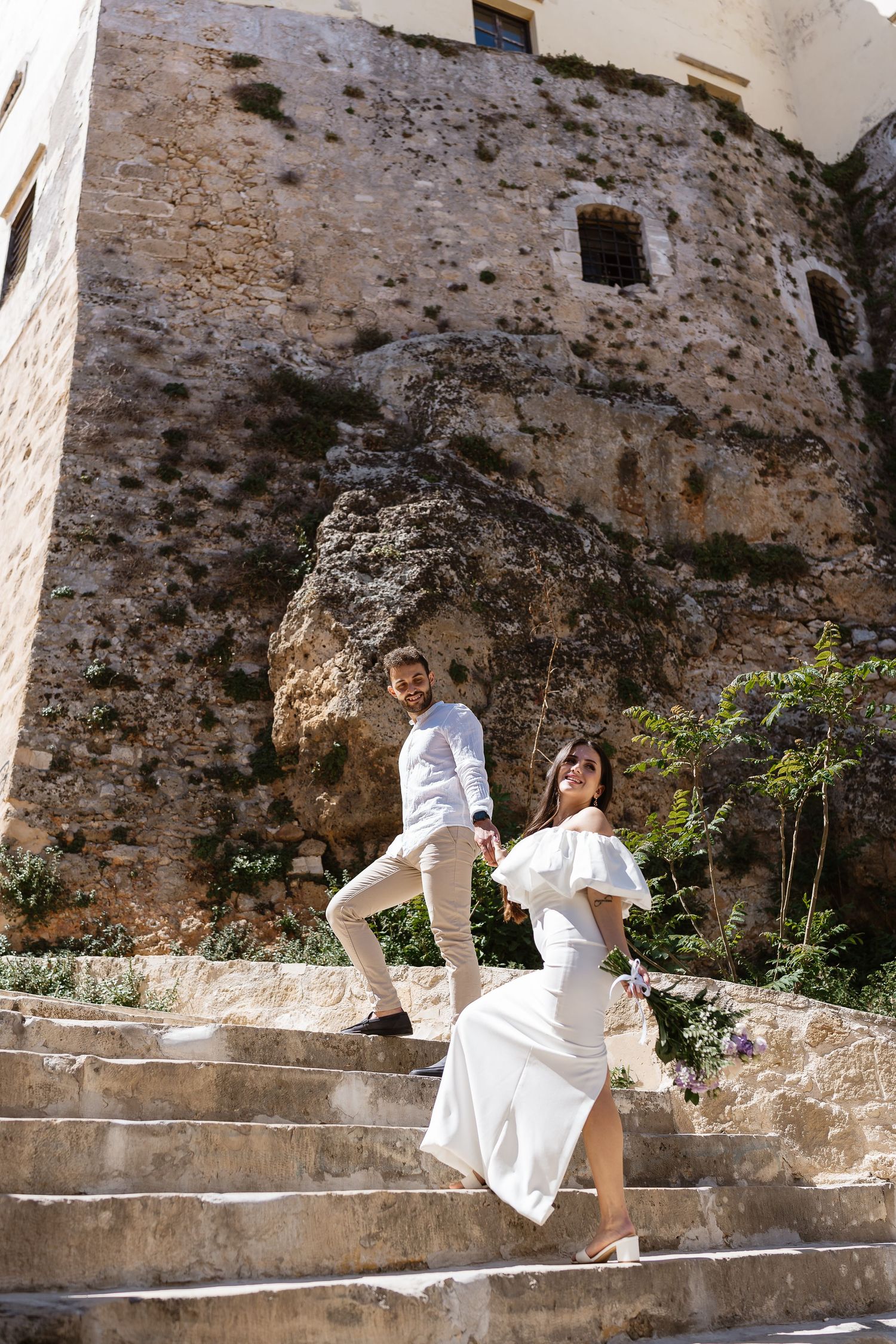 Elegant bride in white off-shoulder dress and groom climbing stone steps beside ancient rock wall.