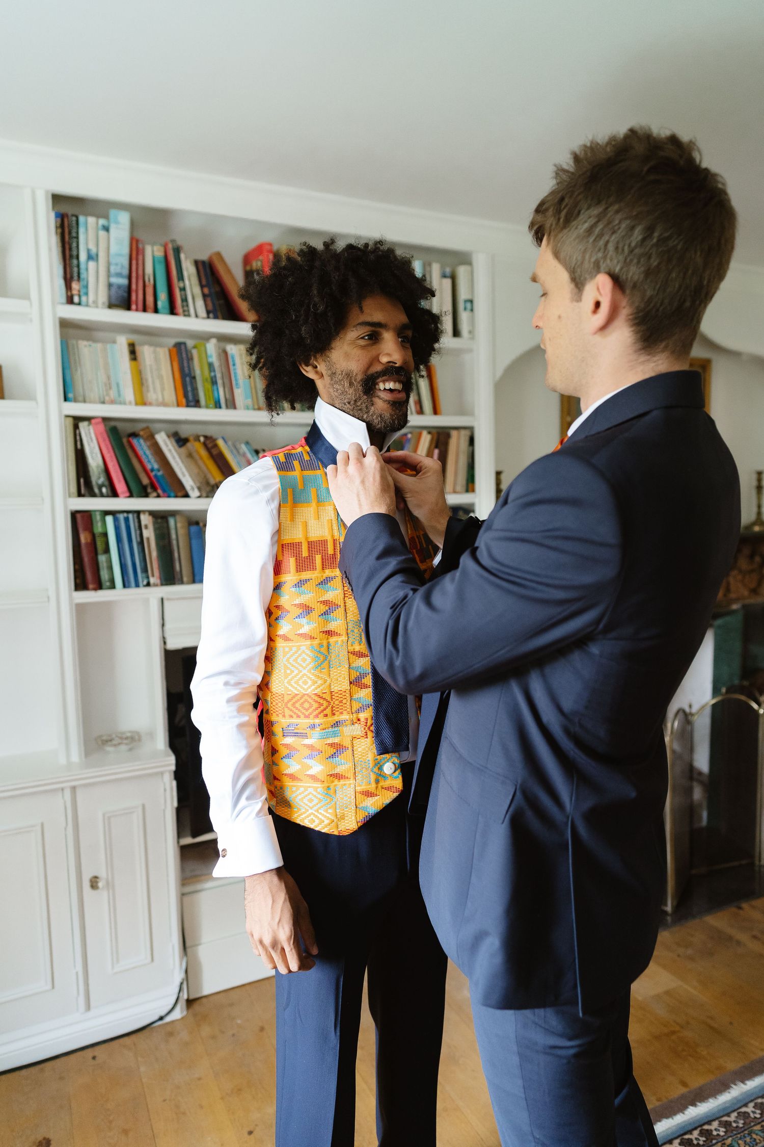 Groomsman helping smiling groom adjust tie while wearing colorful Kente vest indoors.