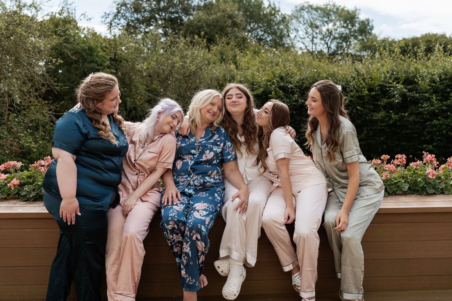 Six women in pastel pajamas laughing and leaning together on a garden wall during bridal party preparations.