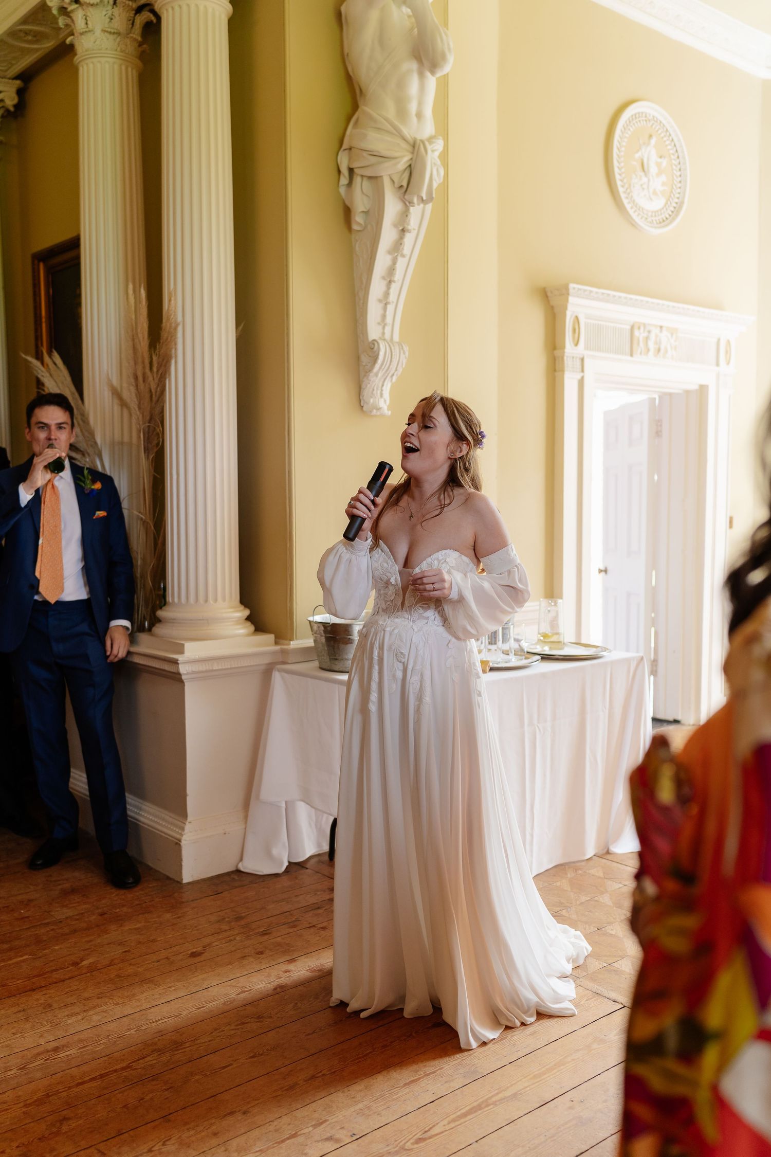Bride sings into microphone in elegant yellow room with white columns during wedding reception.