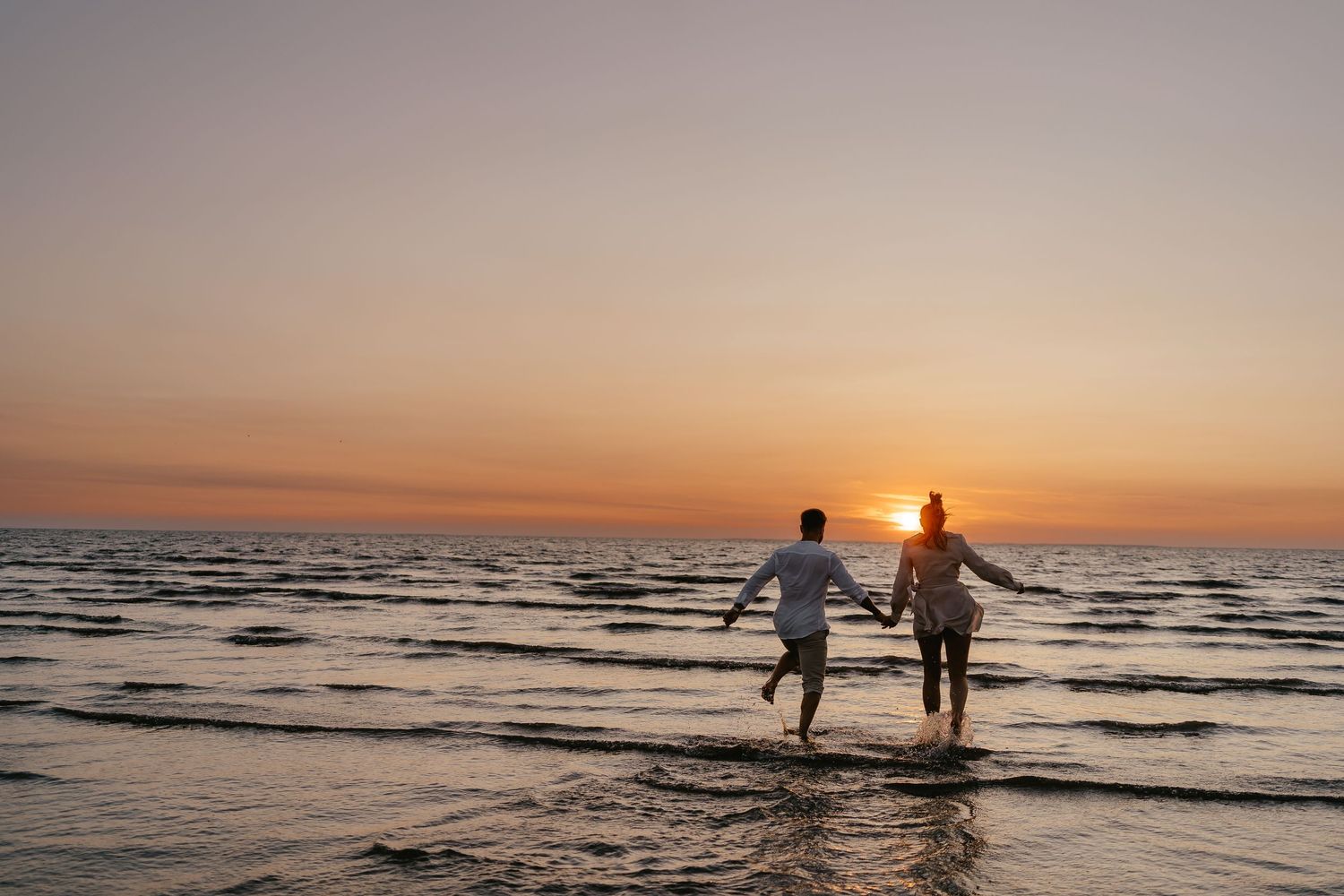 Couple running hand in hand into ocean waves at golden sunset on beach.