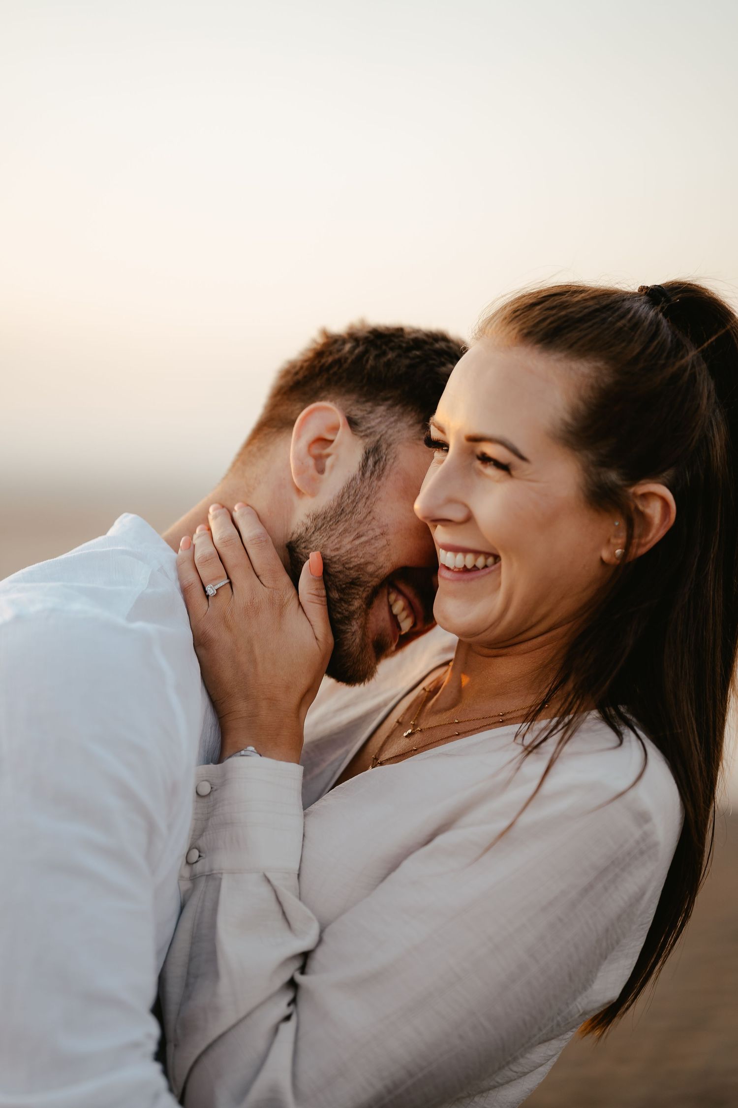 Woman laughing joyfully as man whispers in her ear on a beach at sunset, golden light glowing.