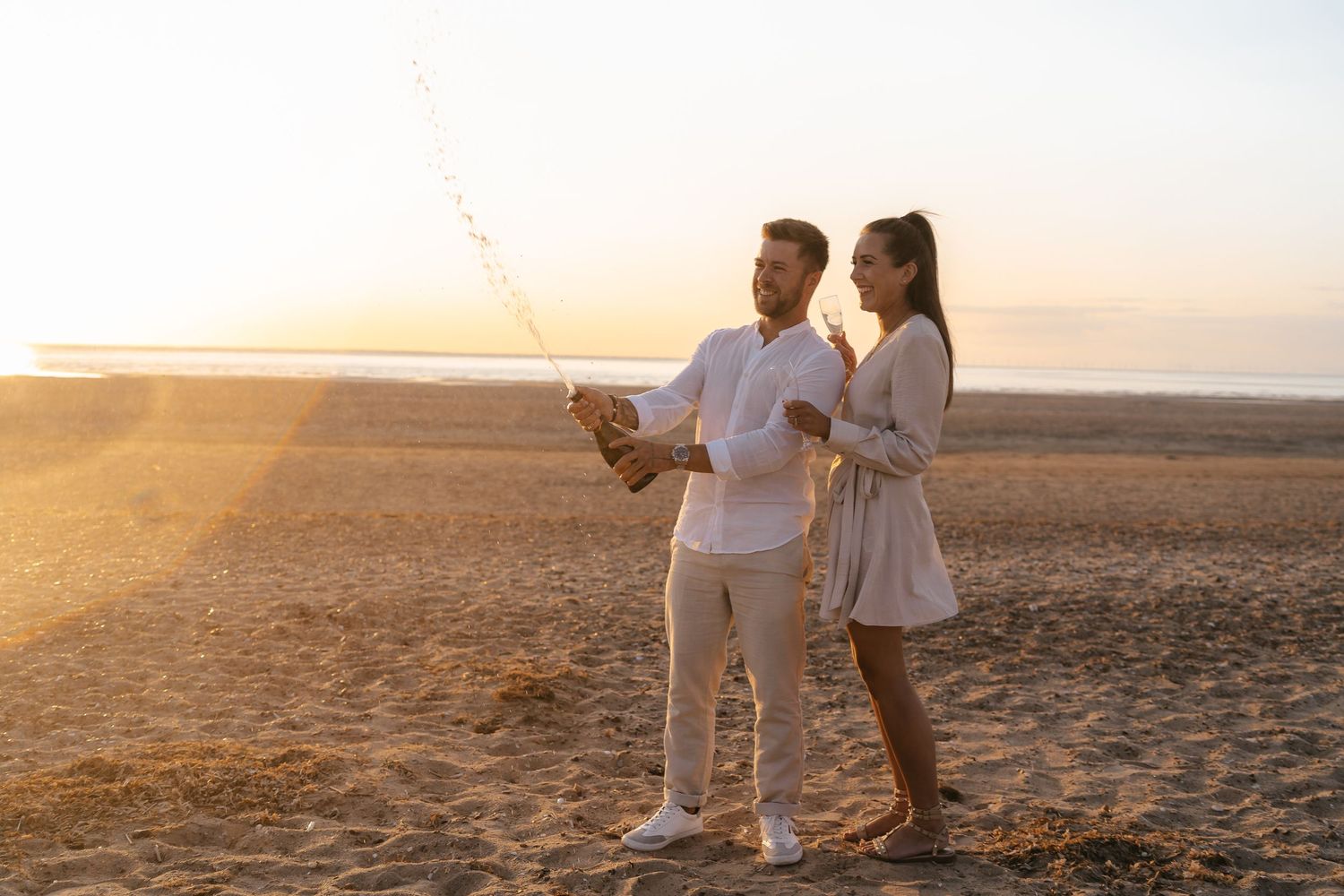 Couple celebrating on a sandy beach at sunset, popping champagne with sparkling stream in the air.