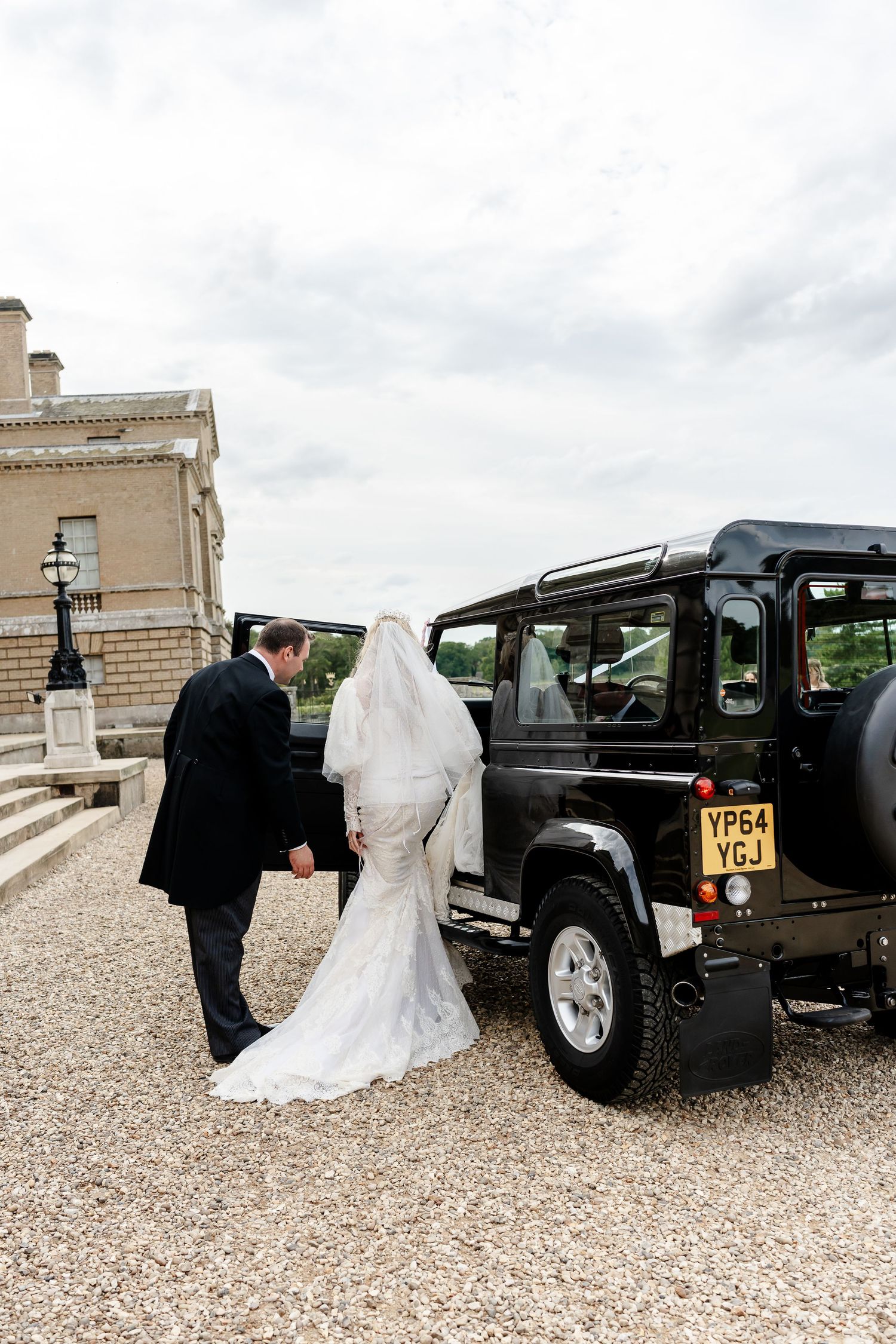 Bride in lace gown and groom approach black Land Rover Defender outside elegant estate.