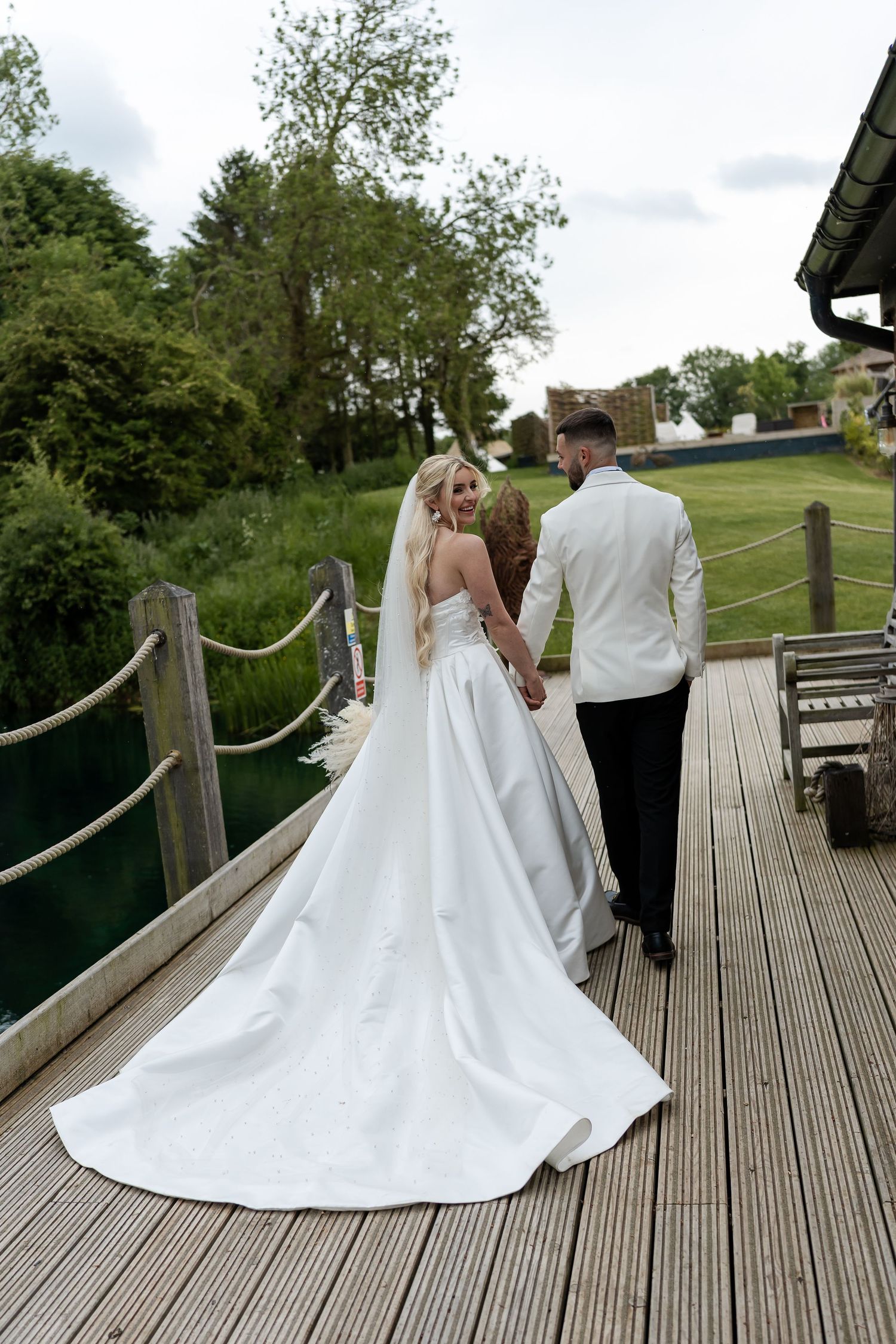 Bride and groom walking hand in hand on a wooden deck bridge surrounded by lush greenery on their wedding day.