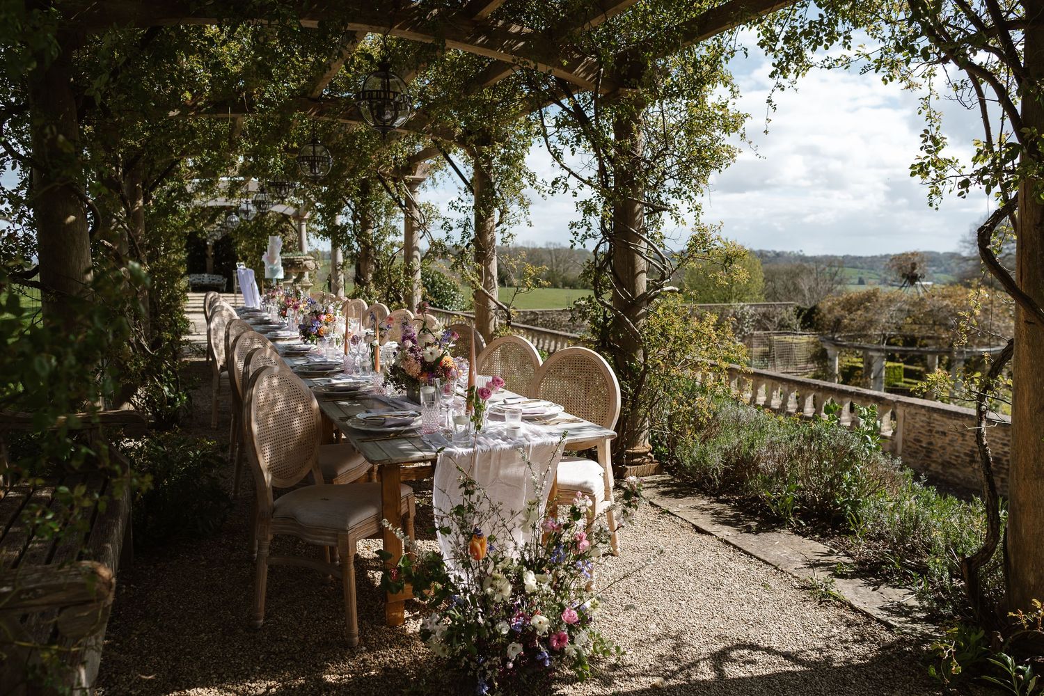 Long outdoor wedding reception table set under vine-covered pergola with countryside views and floral centerpieces.