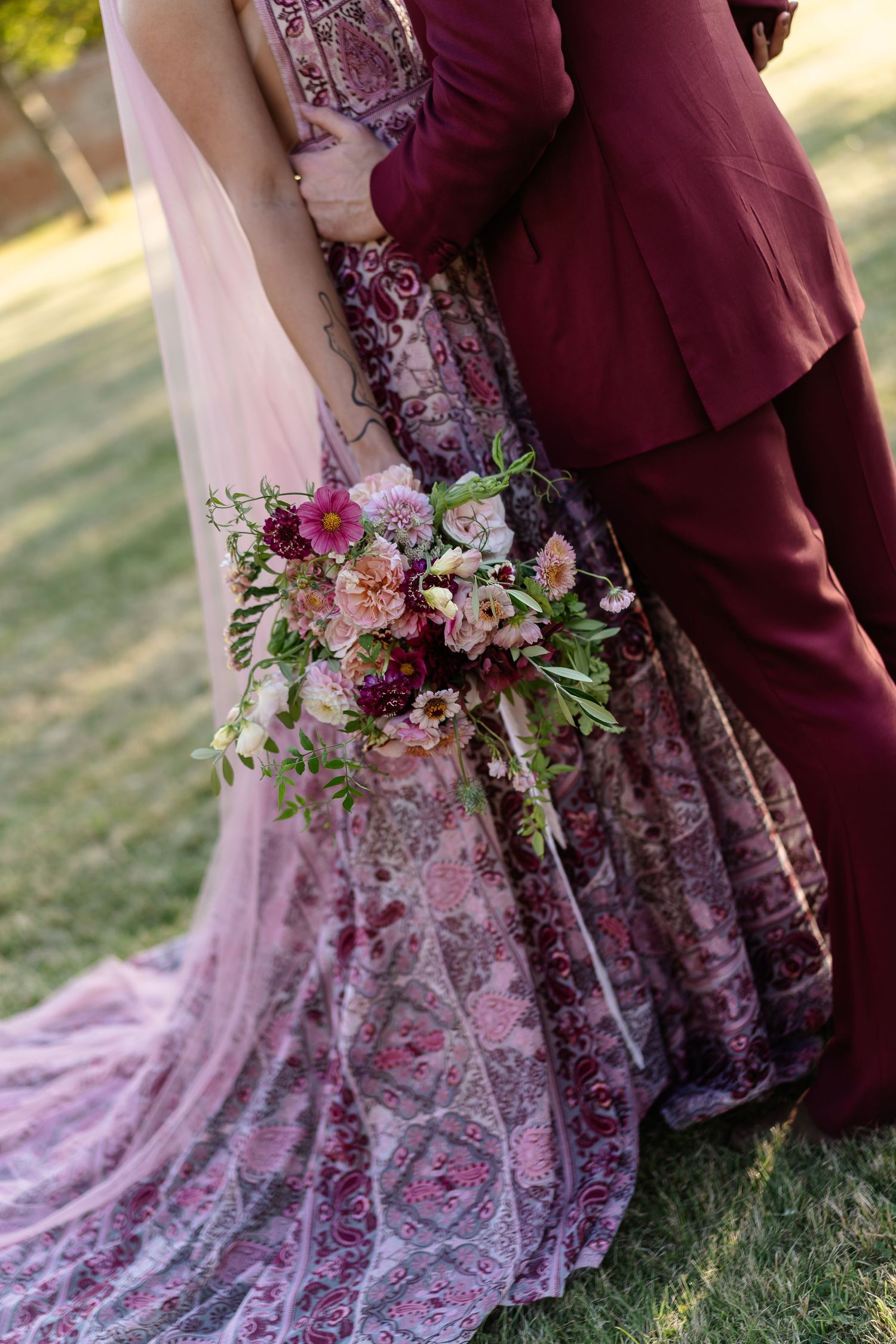 Bride in embroidered pink lehenga holds floral bouquet while embracing groom in burgundy suit.