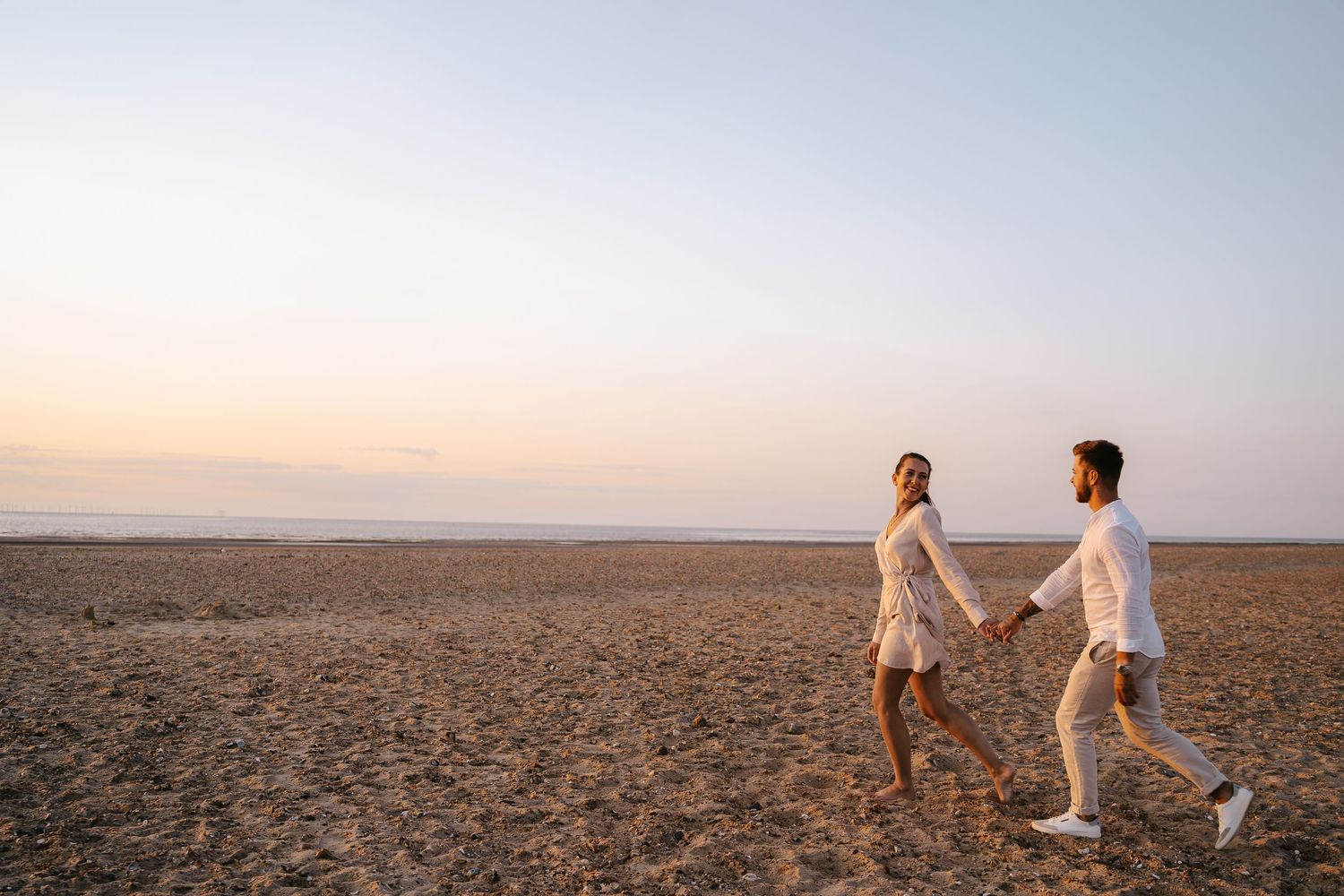 Couple holding hands walking on sandy beach at sunset wearing white outfits.