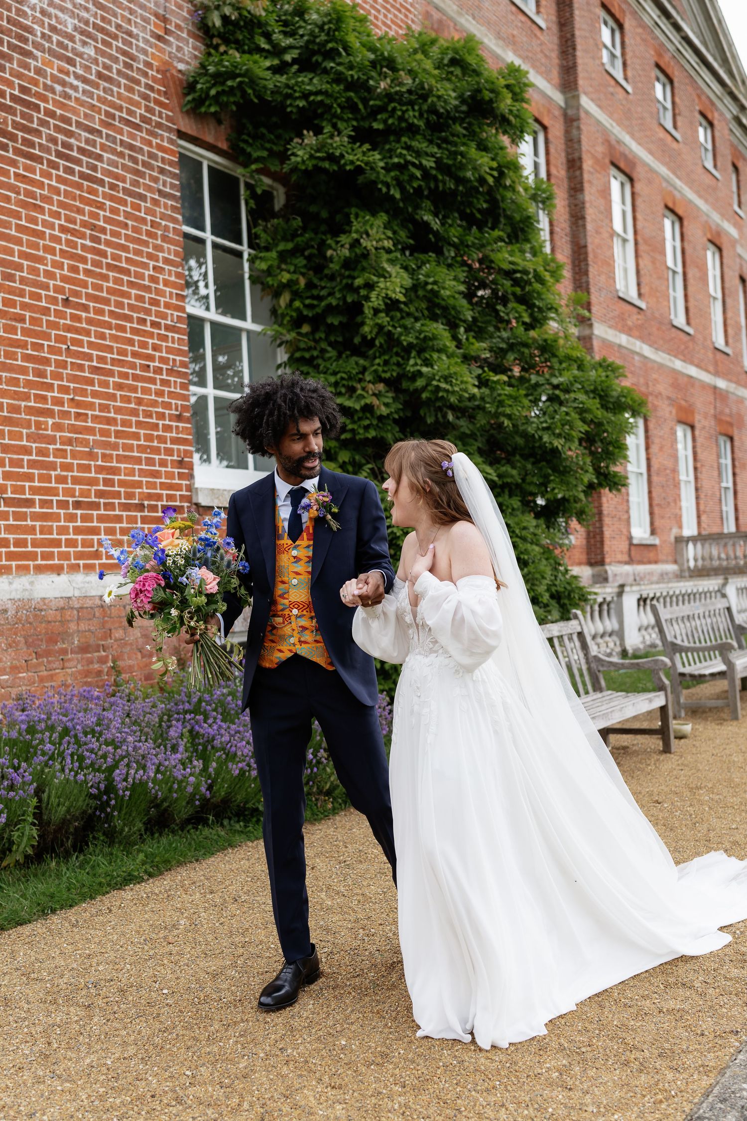 Bride and groom holding hands walking outside a red brick manor house with colorful wildflower bouquet.