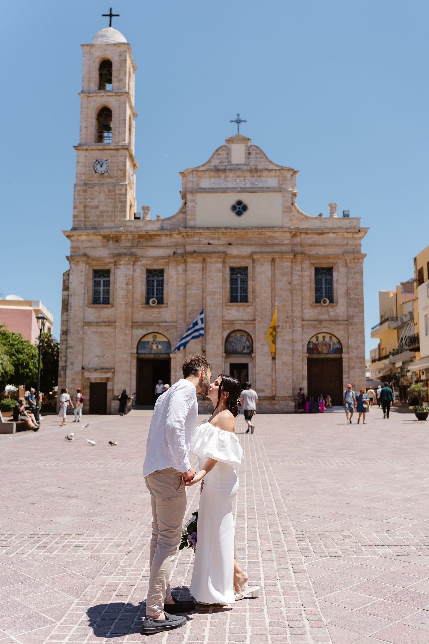Bride and groom kissing in front of a historic church with a Greek flag in Chania, Crete.