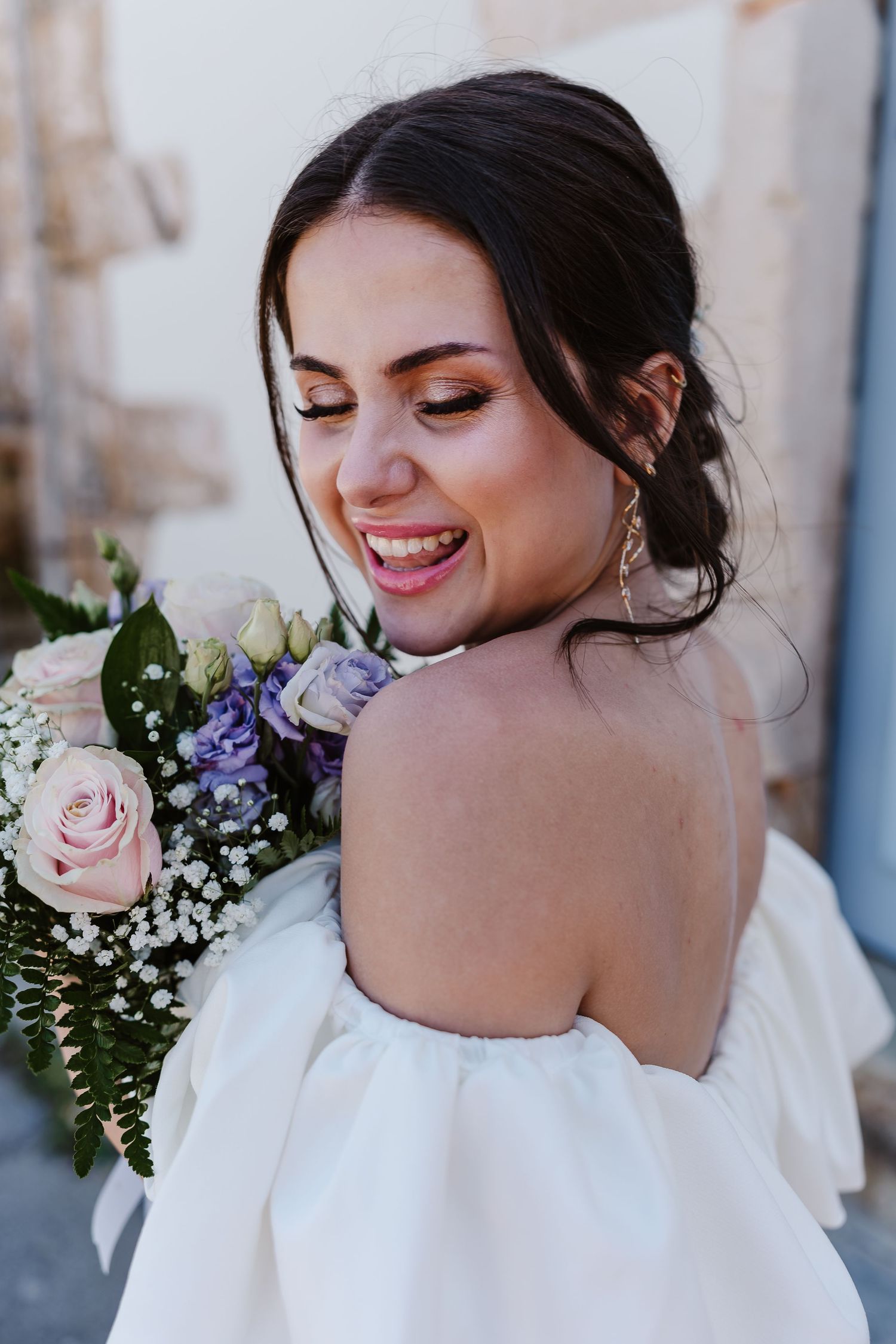 Joyful bride in off-shoulder white dress laughs while holding a pastel floral bouquet outdoors.