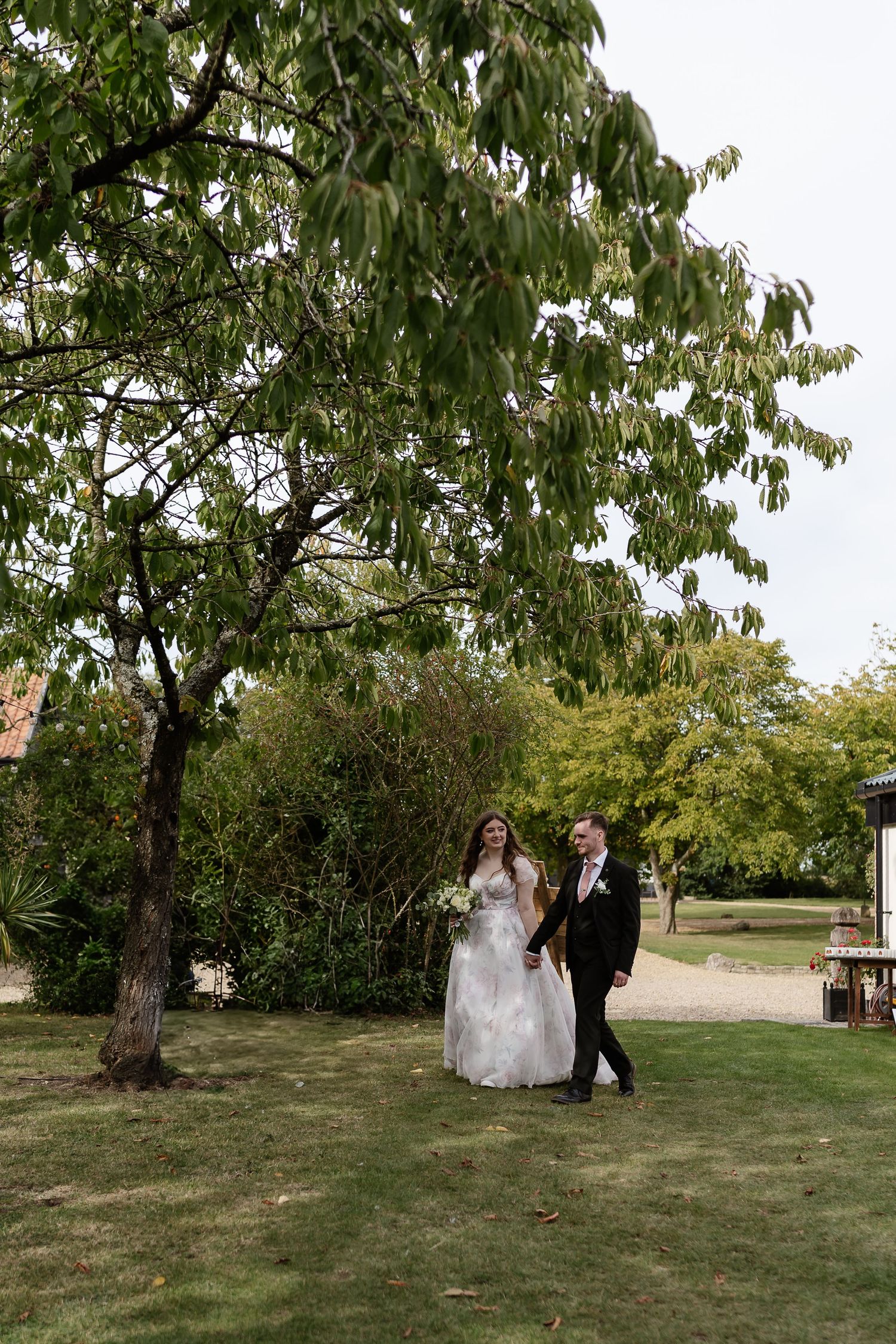 Bride and groom walk hand in hand through a lush garden beneath a large leafy tree.