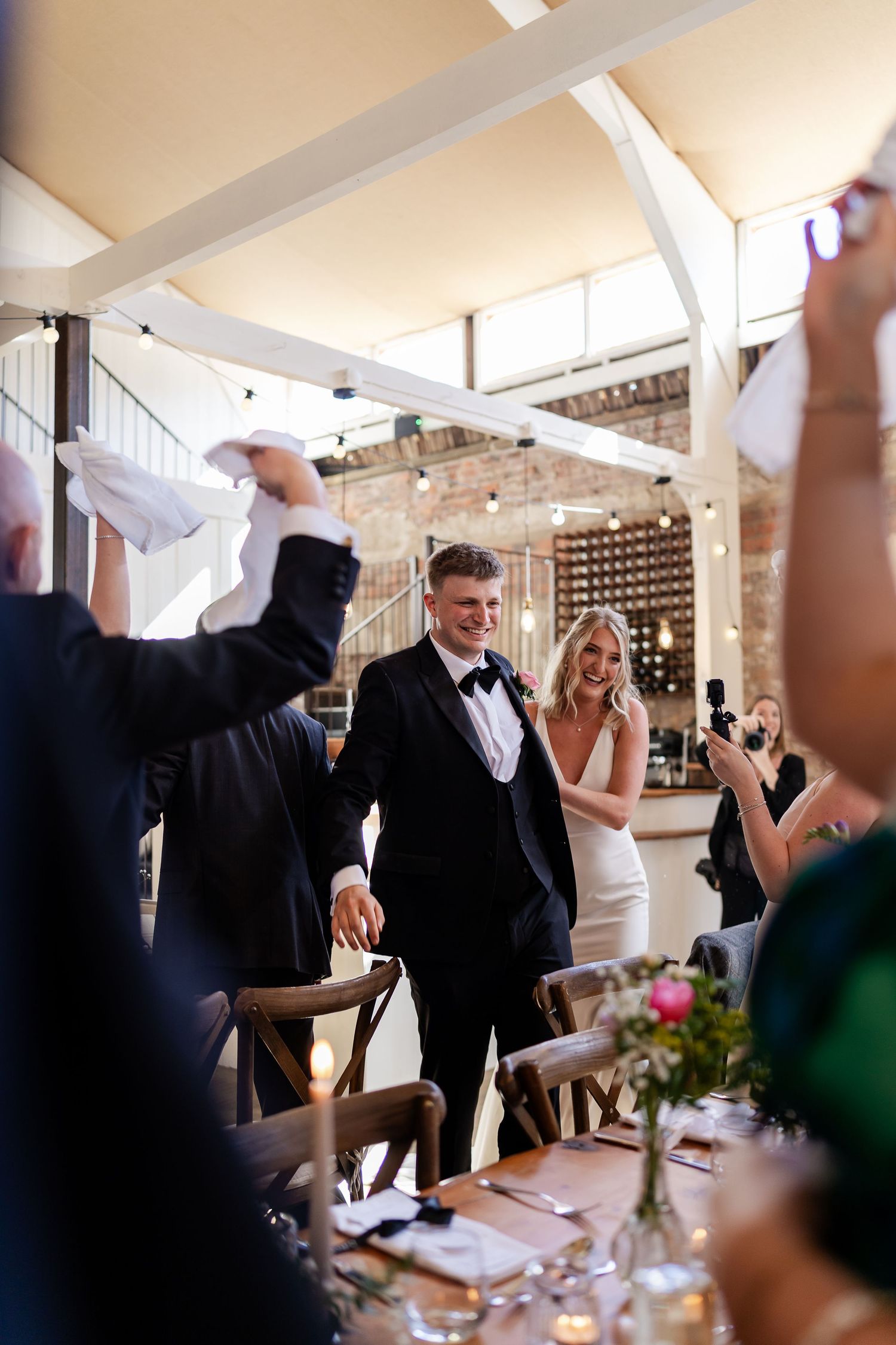 Newlywed couple in formal attire enters wedding reception to cheering guests in rustic barn venue.