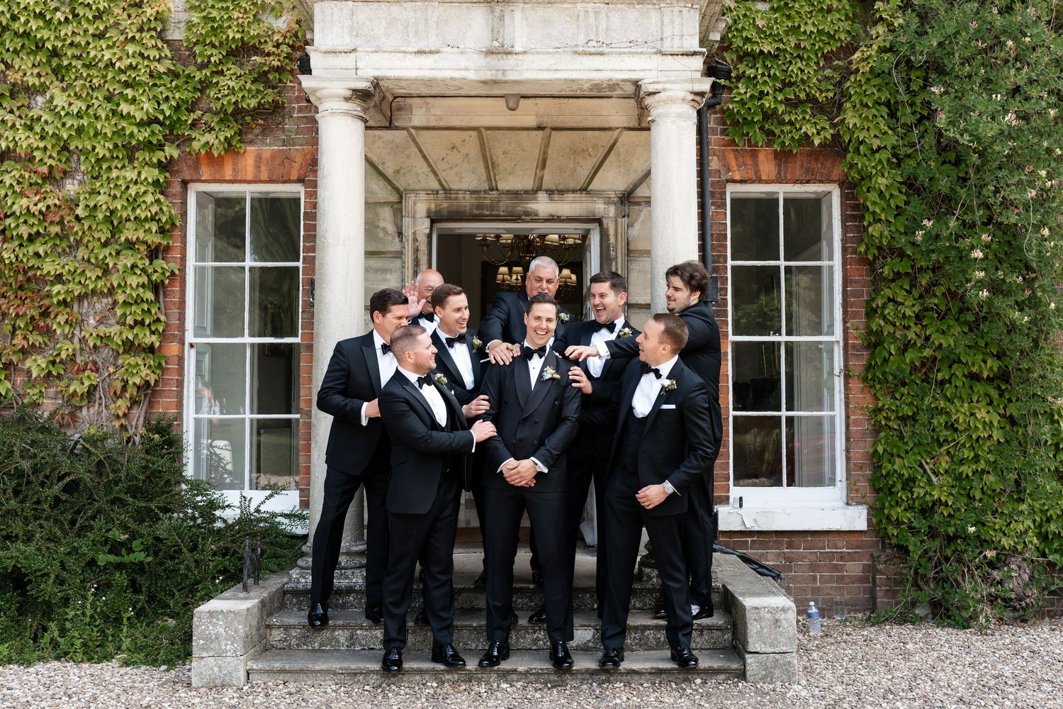 Groomsmen in black tuxedos laughing together on steps of ivy-covered English country house wedding venue.