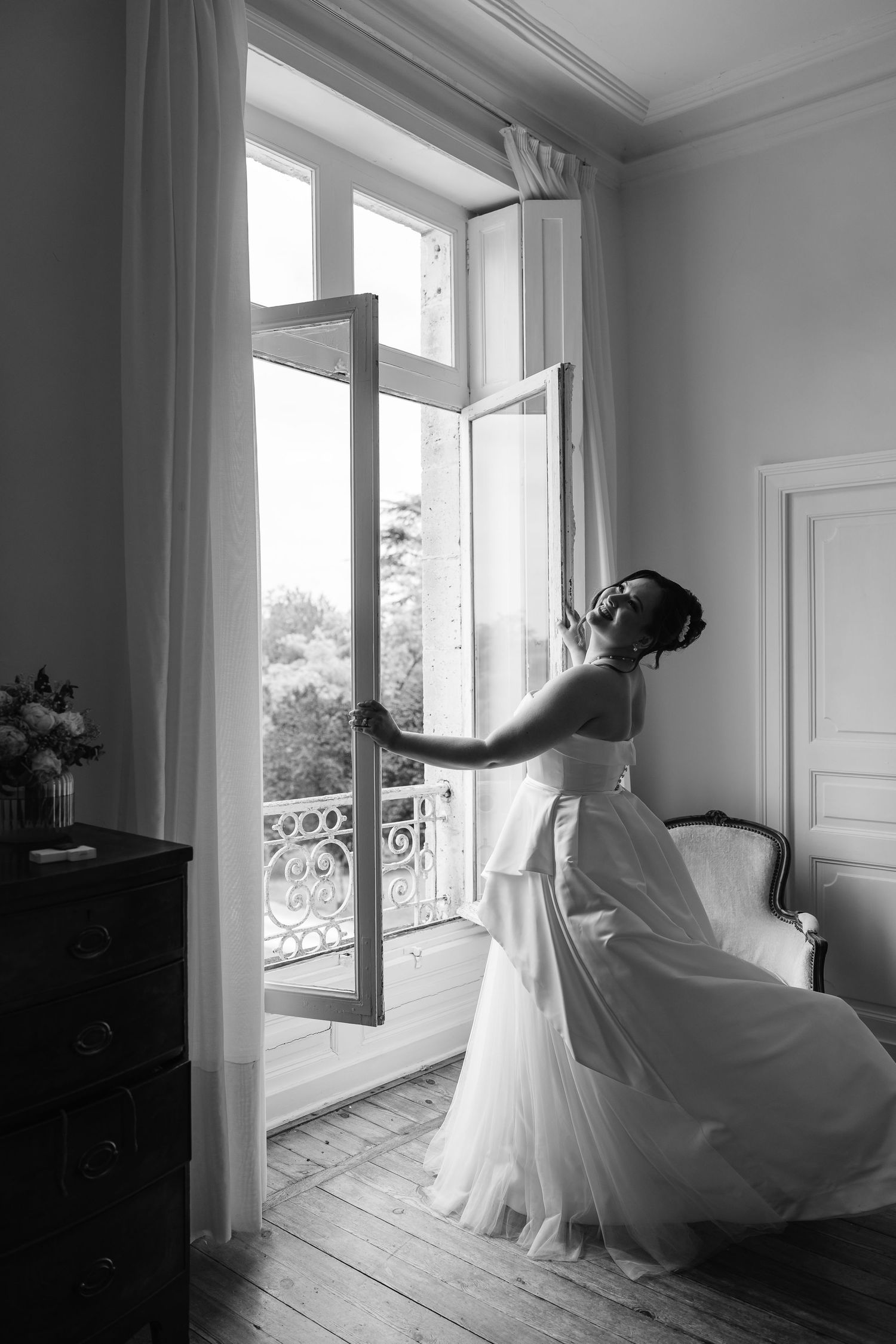 Black and white portrait of bride in strapless gown opening elegant French window in ornate chateau room.