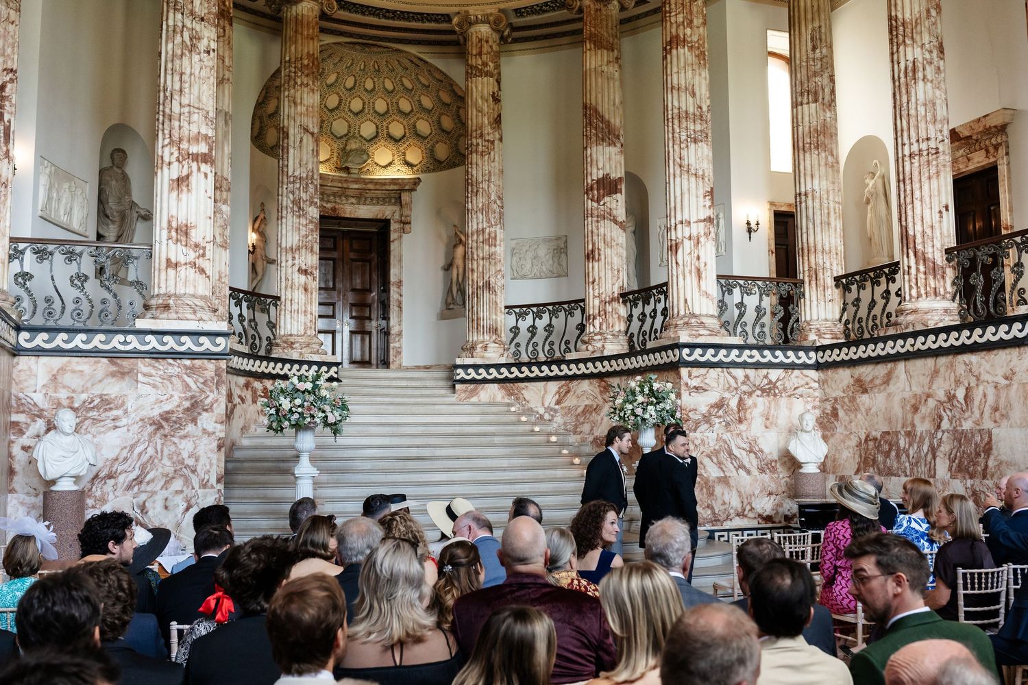Elegant wedding ceremony in a grand marble hall with ornate staircases, floral arrangements, and seated guests.