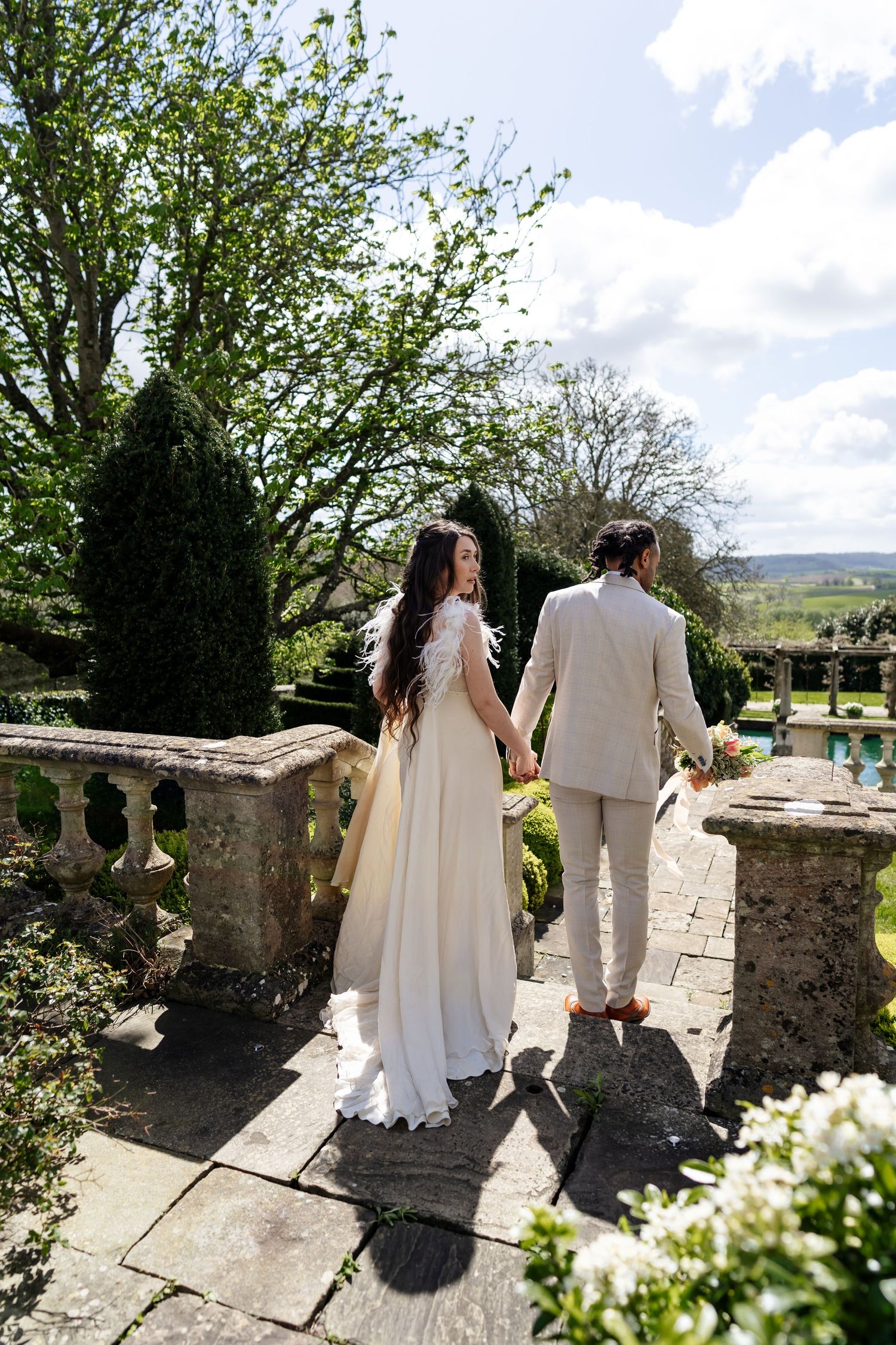 A bride and groom hold hands while walking along a stone balustrade in a formal garden.