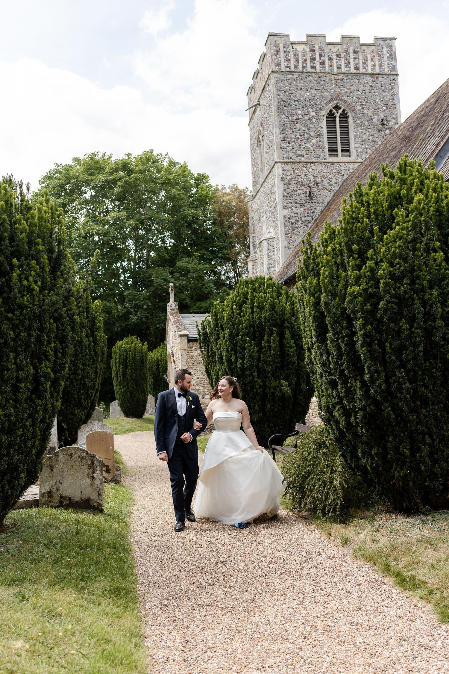 Bride and groom walking along gravel path lined with tall hedges beside historic stone church tower.
