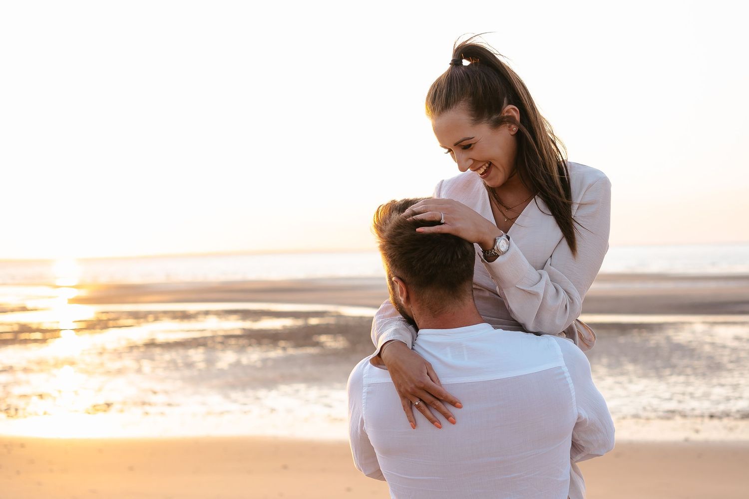 Romantic couple embracing on beach at sunset, woman smiling while being held up.