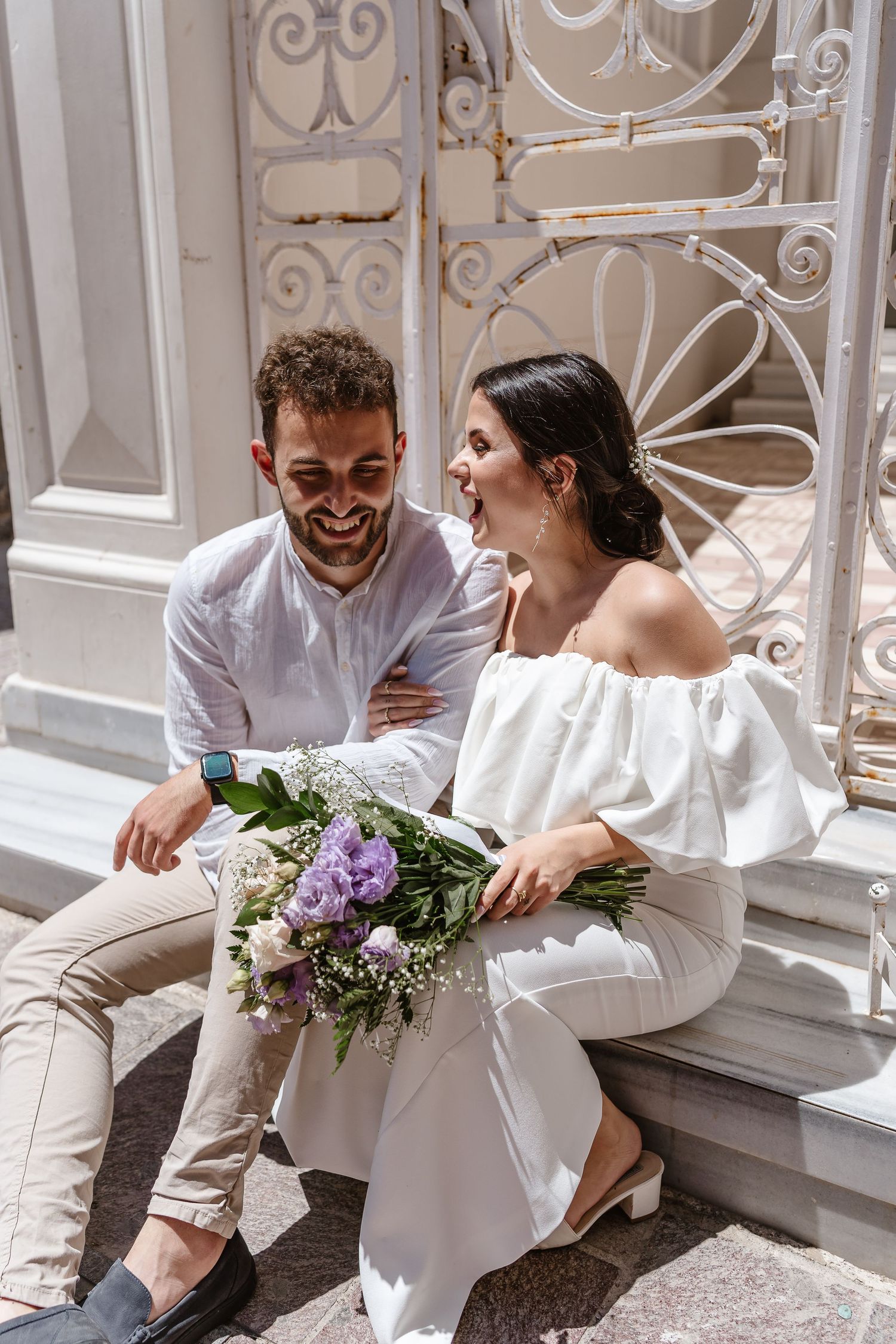 Happy couple in white sitting on steps, bride holding purple and white bouquet, laughing together near ornate gate.