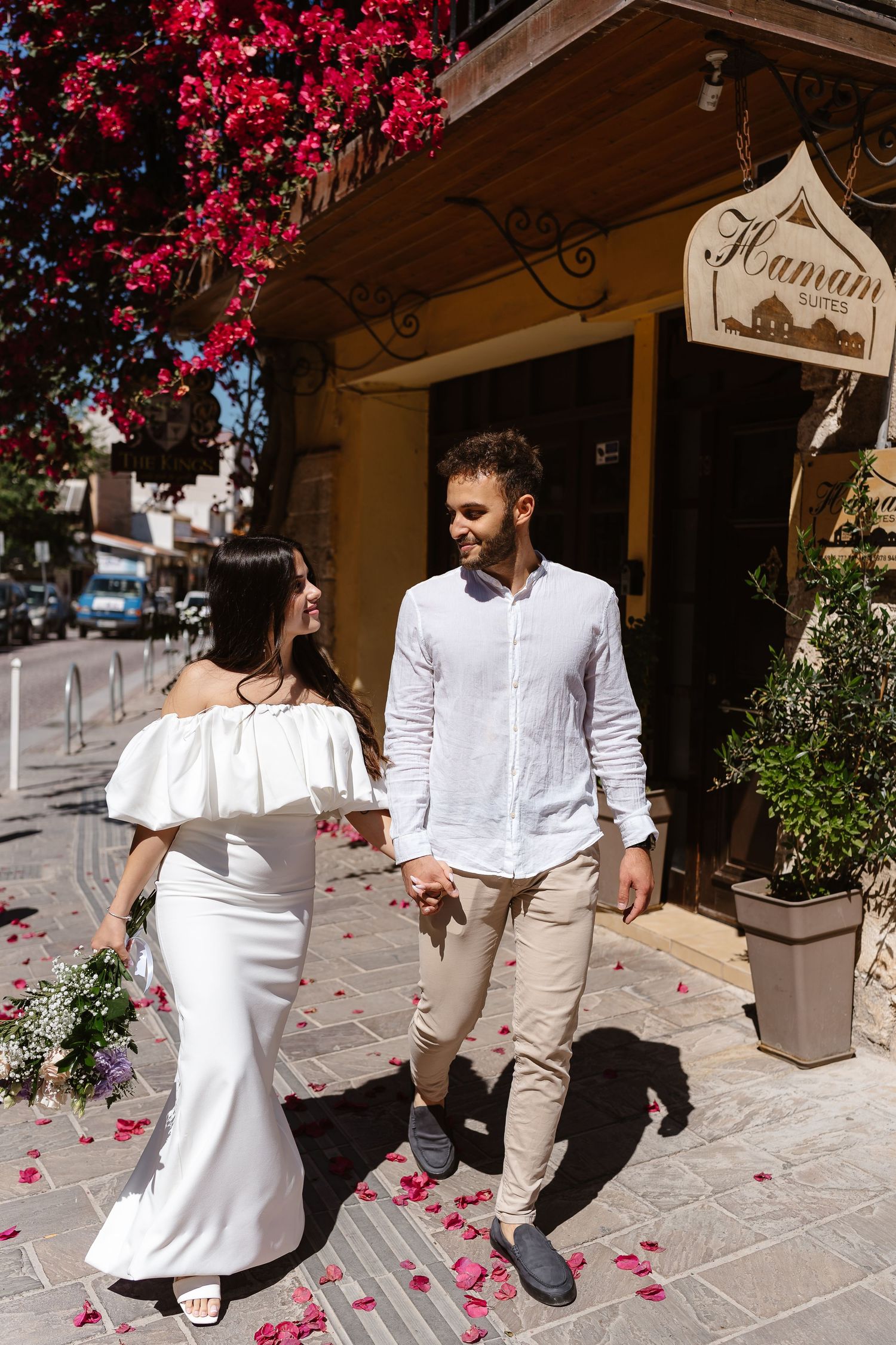Smiling bride and groom holding hands outside a hotel entrance adorned with red flowering bougainvillea.
