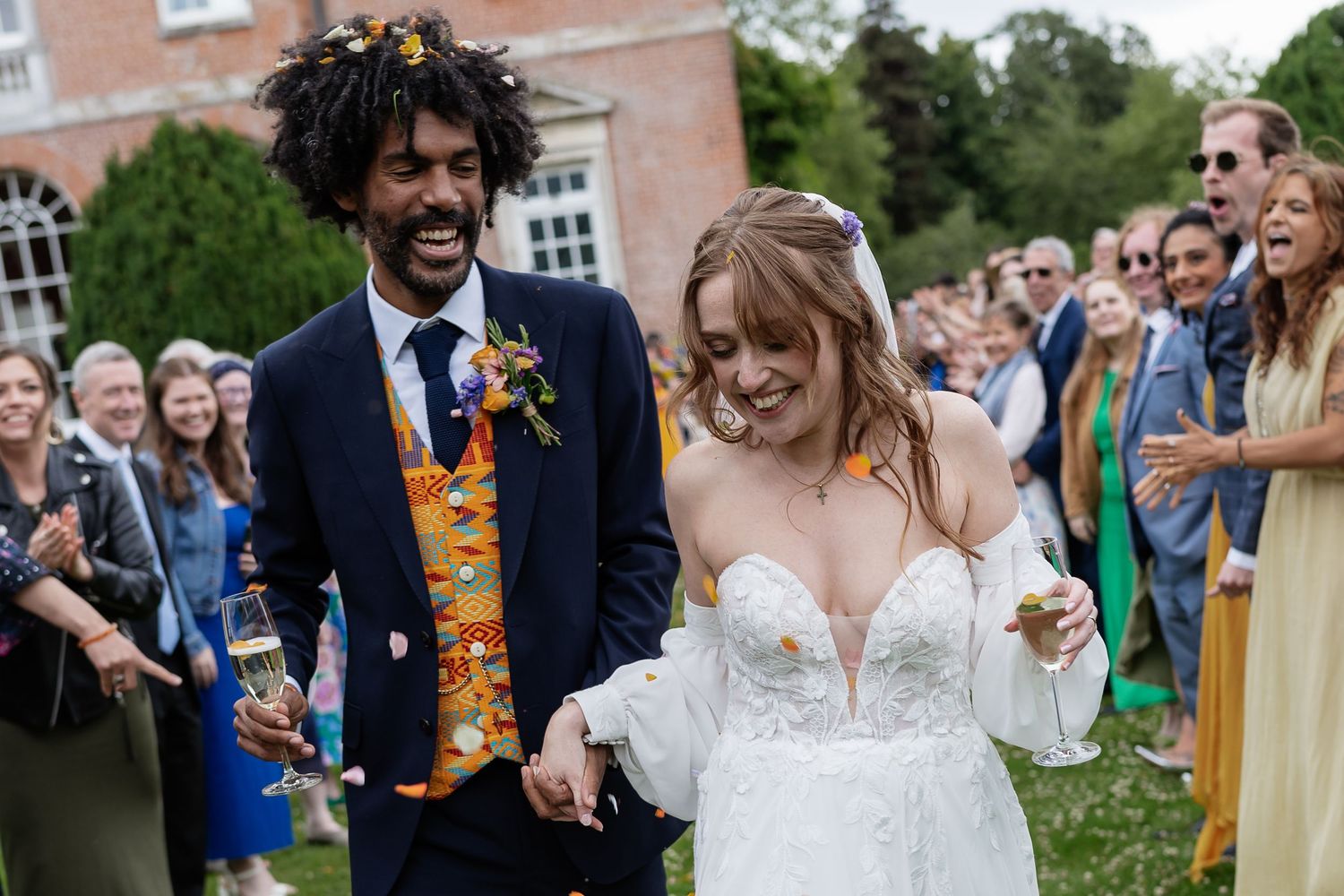 Bride and groom smile holding hands during outdoor confetti celebration with colorful wedding guests.