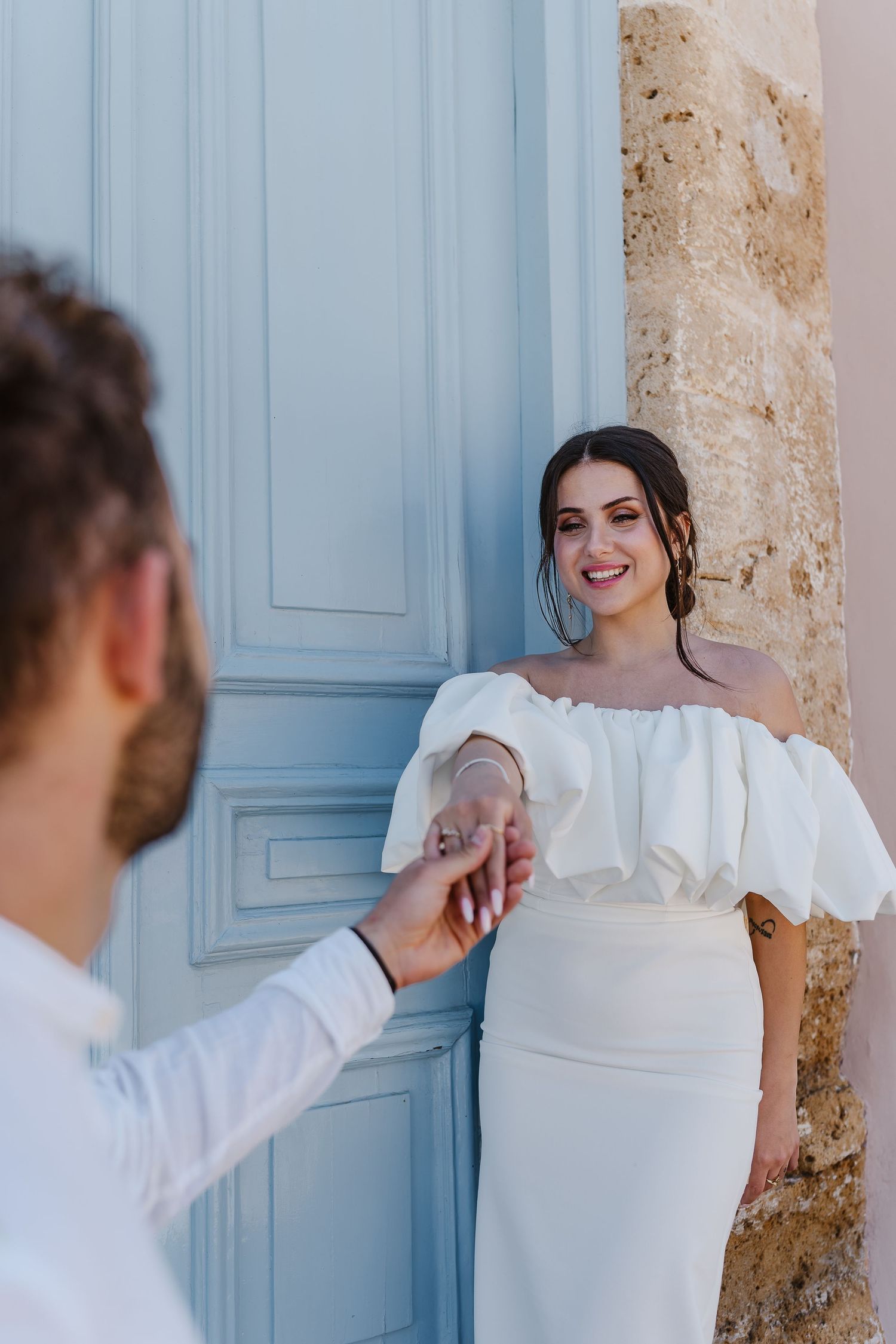 Smiling bride in ruffled off-shoulder white gown holds grooms hand near a light blue door.
