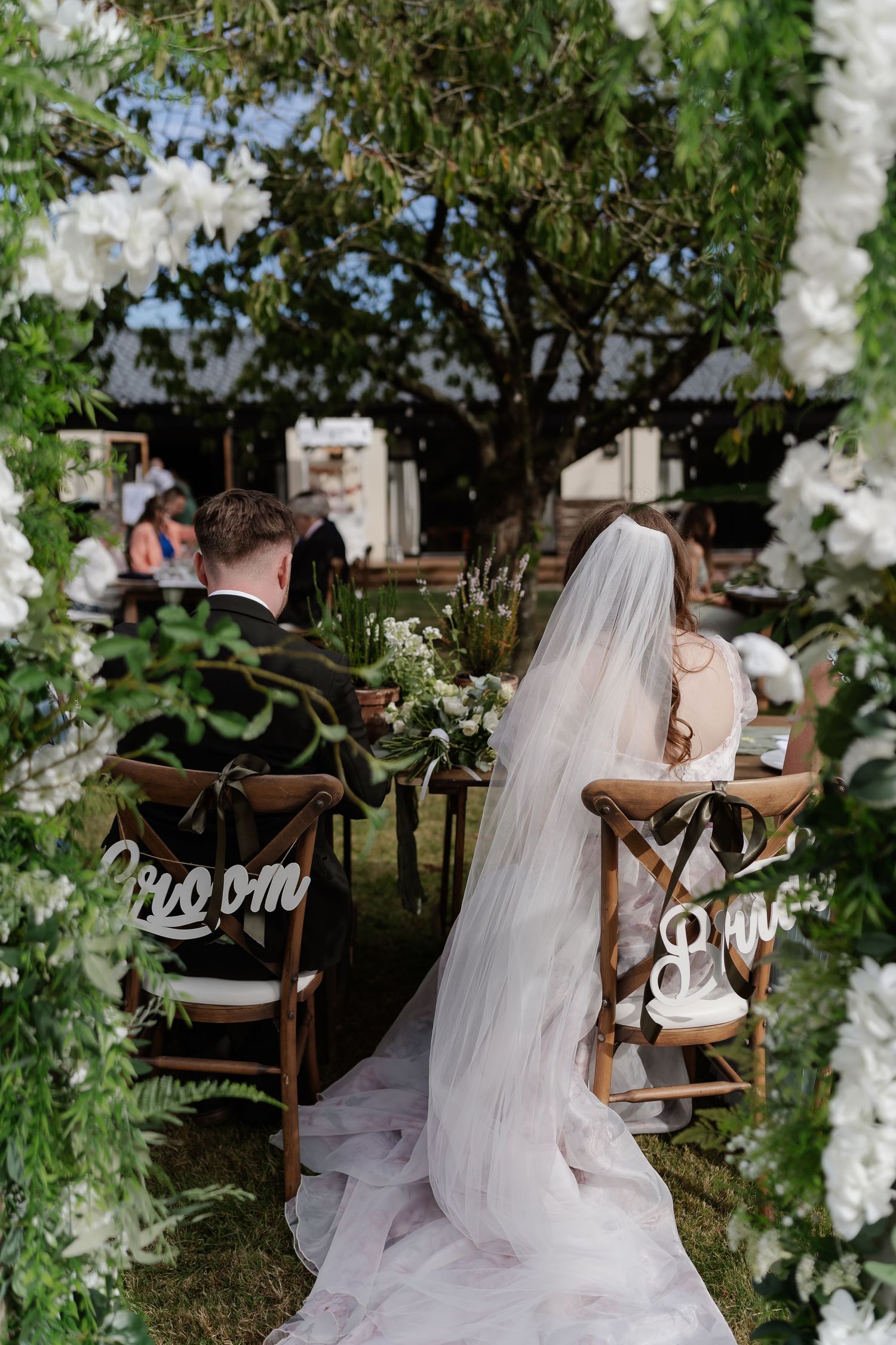 Bride and groom seated at outdoor wedding ceremony altar adorned with white flowers and lush greenery.