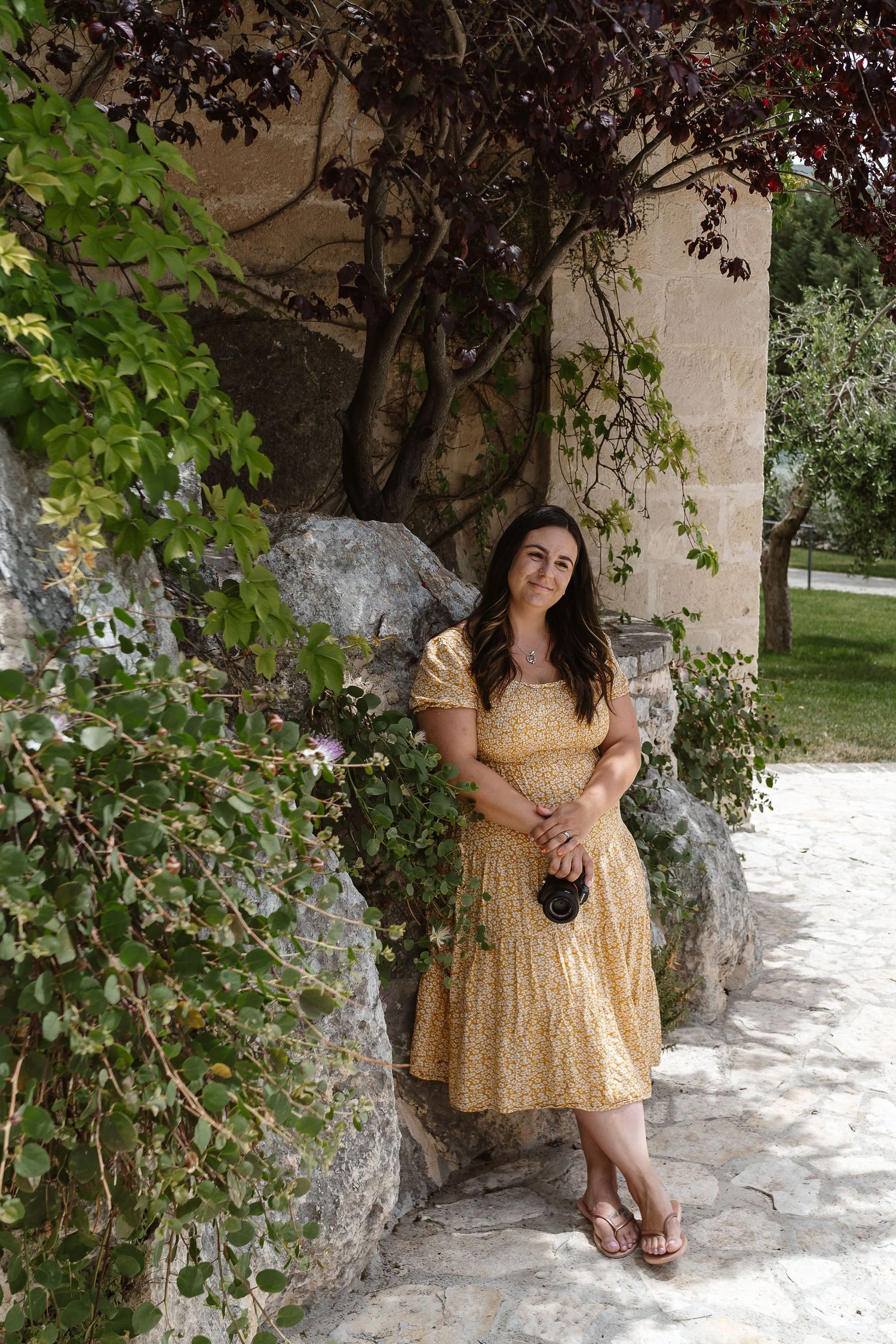 Woman in yellow floral midi dress leaning against stone wall surrounded by lush green ivy in outdoor garden.