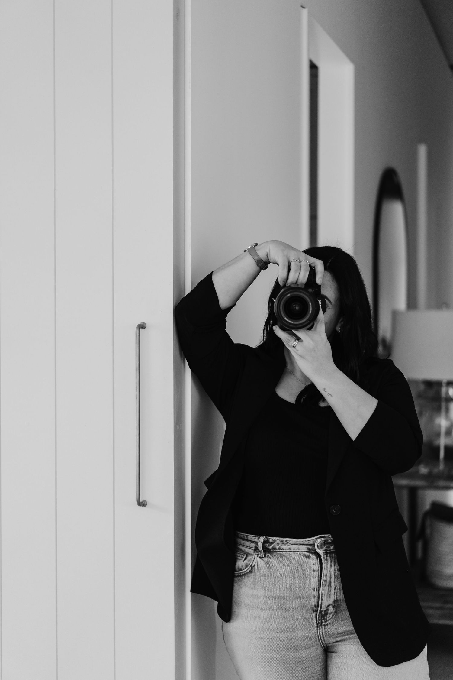 Black and white photo of woman in black blazer photographing with DSLR camera raised to her face indoors.