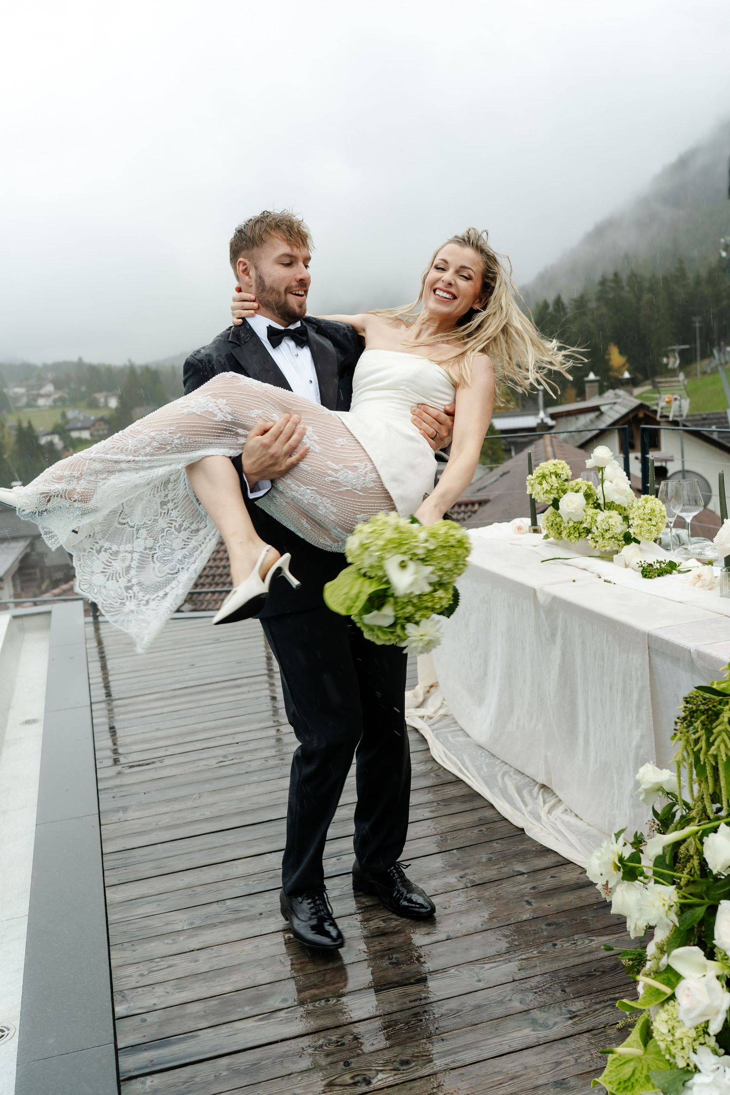 Groom in tuxedo lifts laughing bride on a rainy rooftop terrace with misty mountain views behind them.