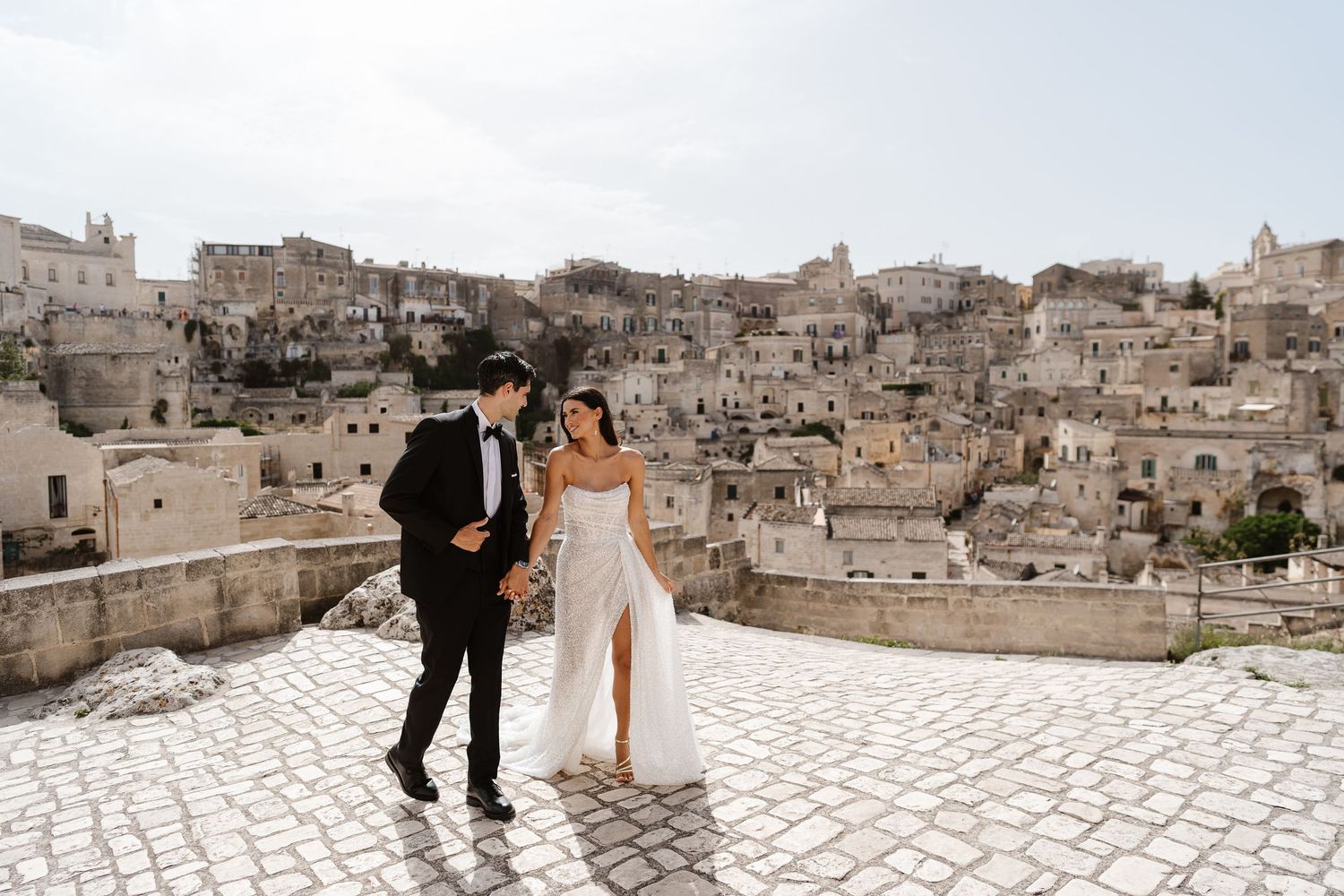 Elegant bride and groom walking on rooftop terrace with stunning ancient Matera cityscape backdrop.