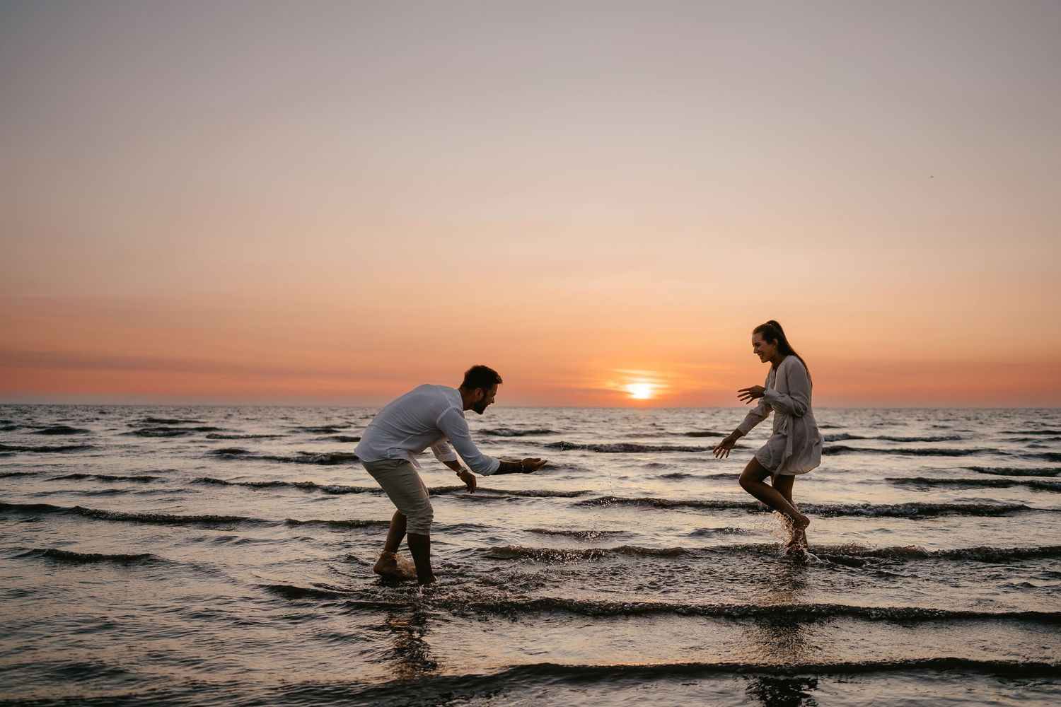 Playful couple splashing water at each other on beach during vibrant sunset.