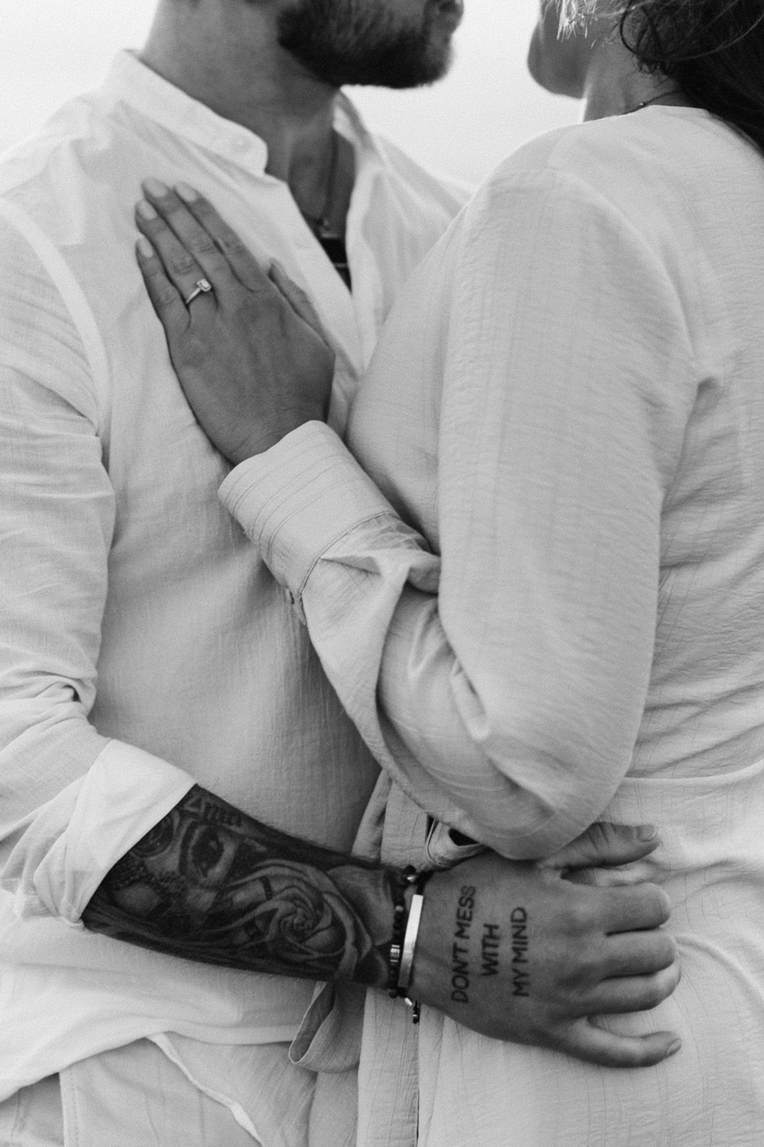 Black and white photo of a couple embracing, showing tattooed arms, a ring, and matching white linen outfits.