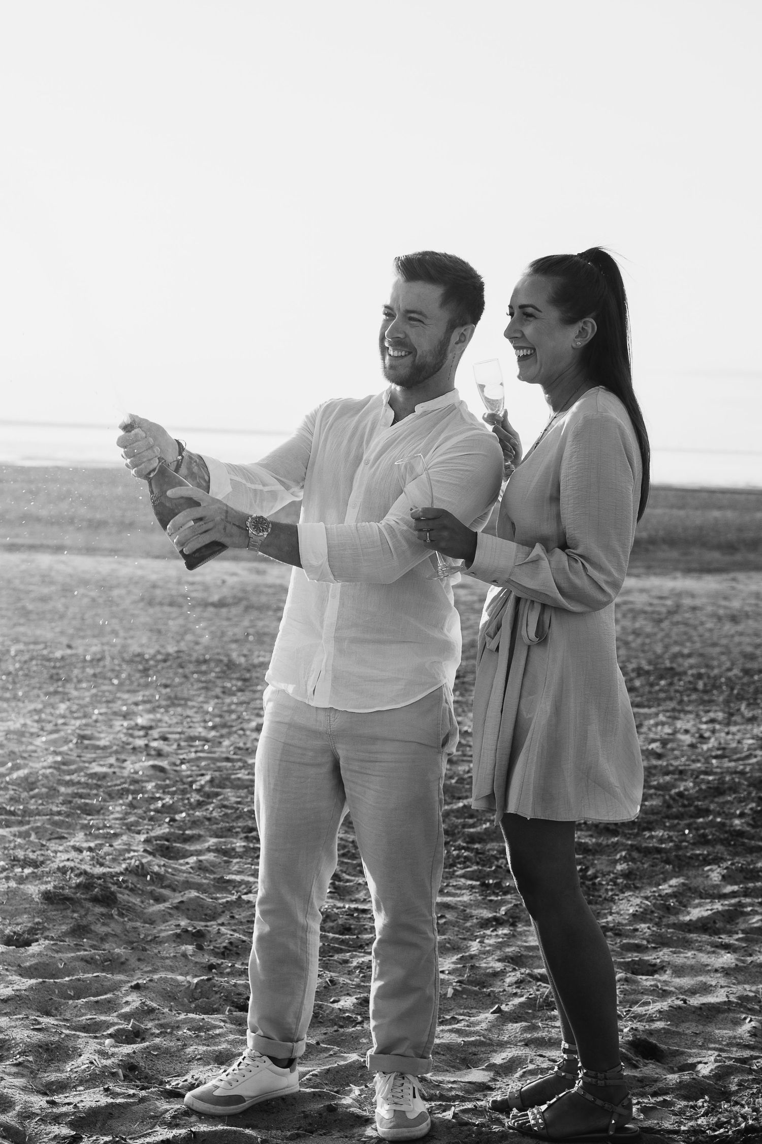 Black and white photo of a joyful couple popping champagne on a beach at sunset.