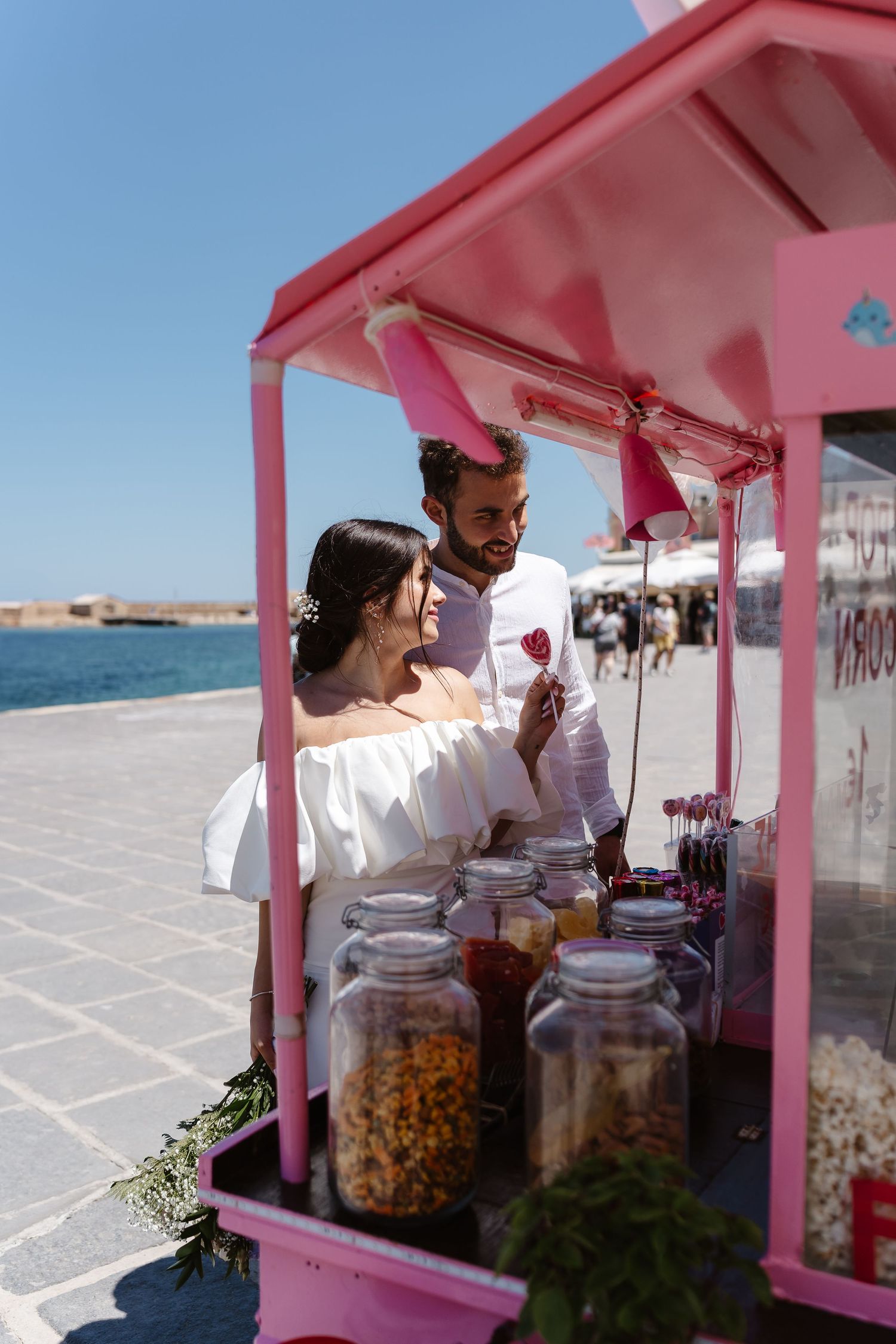 Bride holding a lollipop smiling at groom beside a pink popcorn cart with ocean views in background.