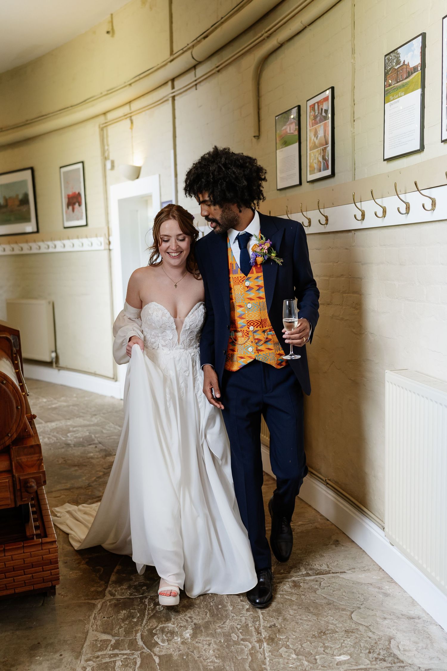 Bride and groom walking together indoors, she in a white gown, he in a navy suit with colorful kente vest holding champagne.