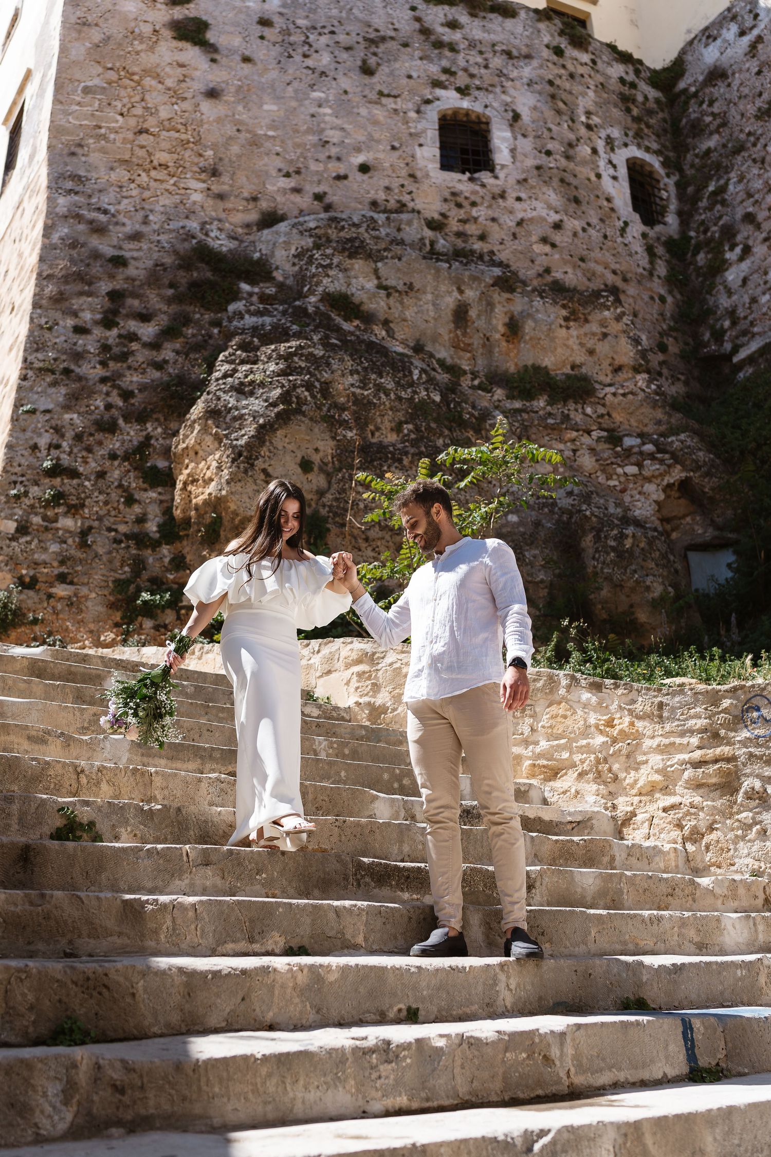 Bride and groom holding hands walking down ancient stone steps at a historic Mediterranean venue.