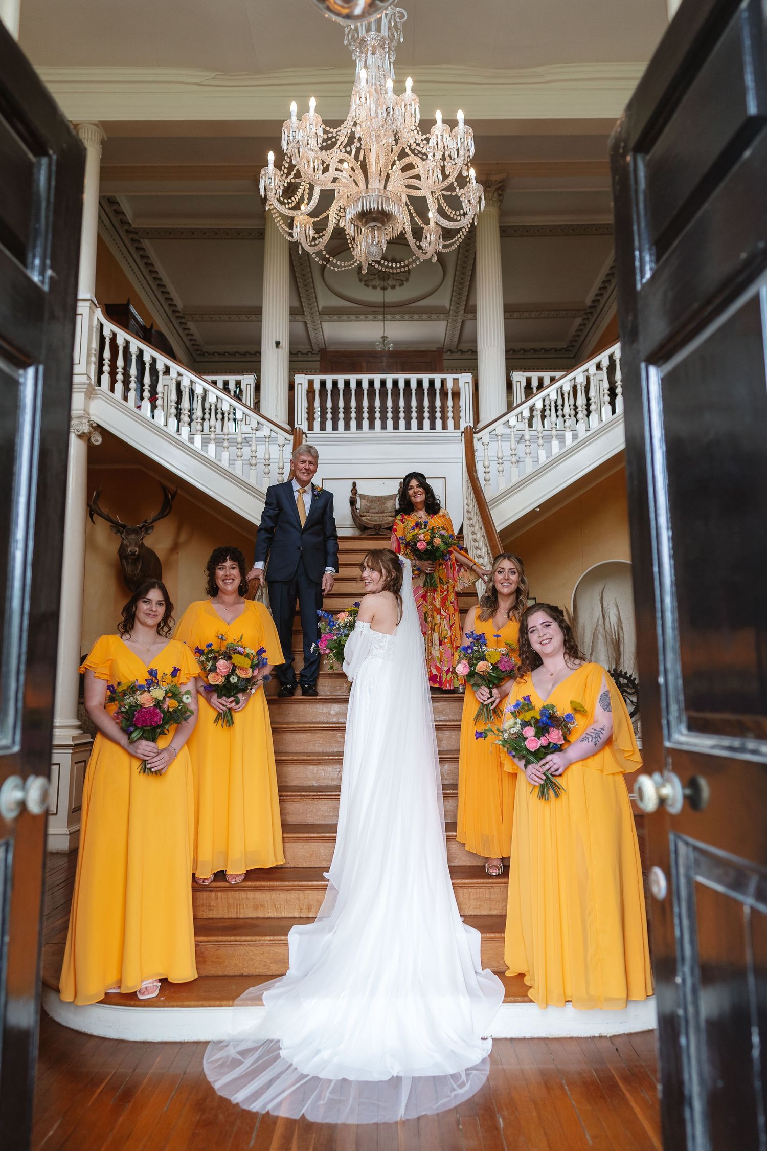 Bride with bridesmaids in yellow dresses descending grand staircase beneath crystal chandelier in manor.