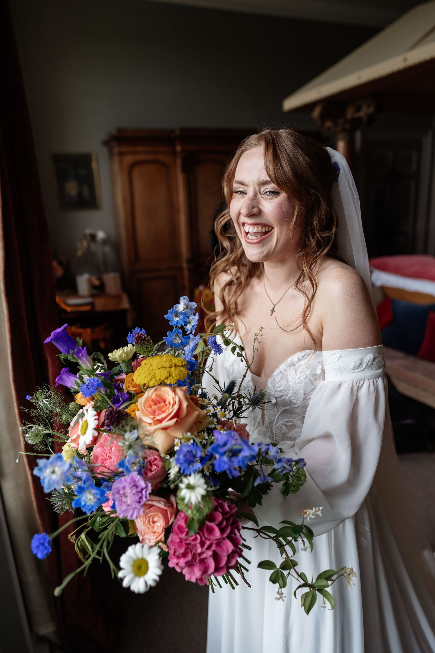 Laughing bride in white off-shoulder gown holding vibrant colorful wedding bouquet indoors.