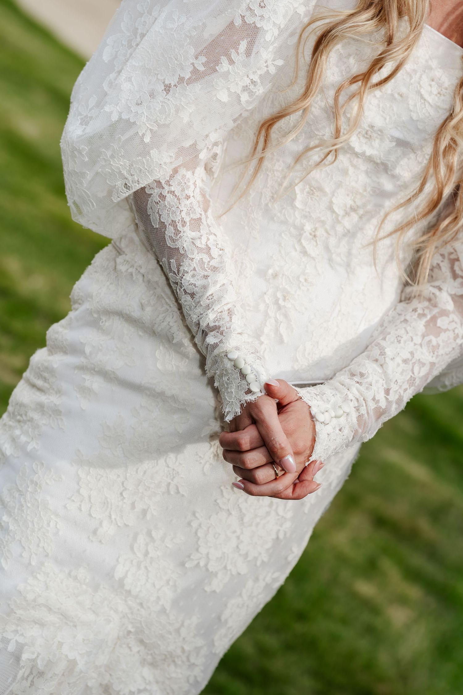 Close-up of bride in long-sleeve lace wedding gown with clasped hands on green lawn.