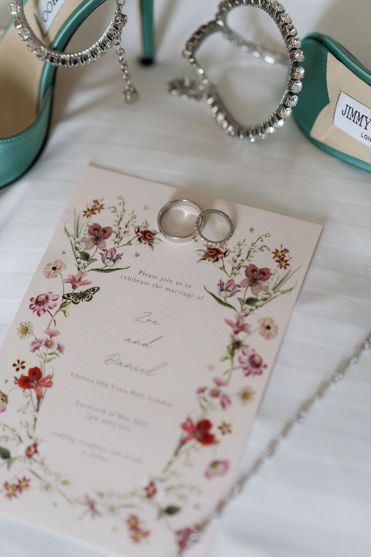 Floral wedding invitation for Zoe and Daniel with rings and teal Jimmy Choo heels on white surface.