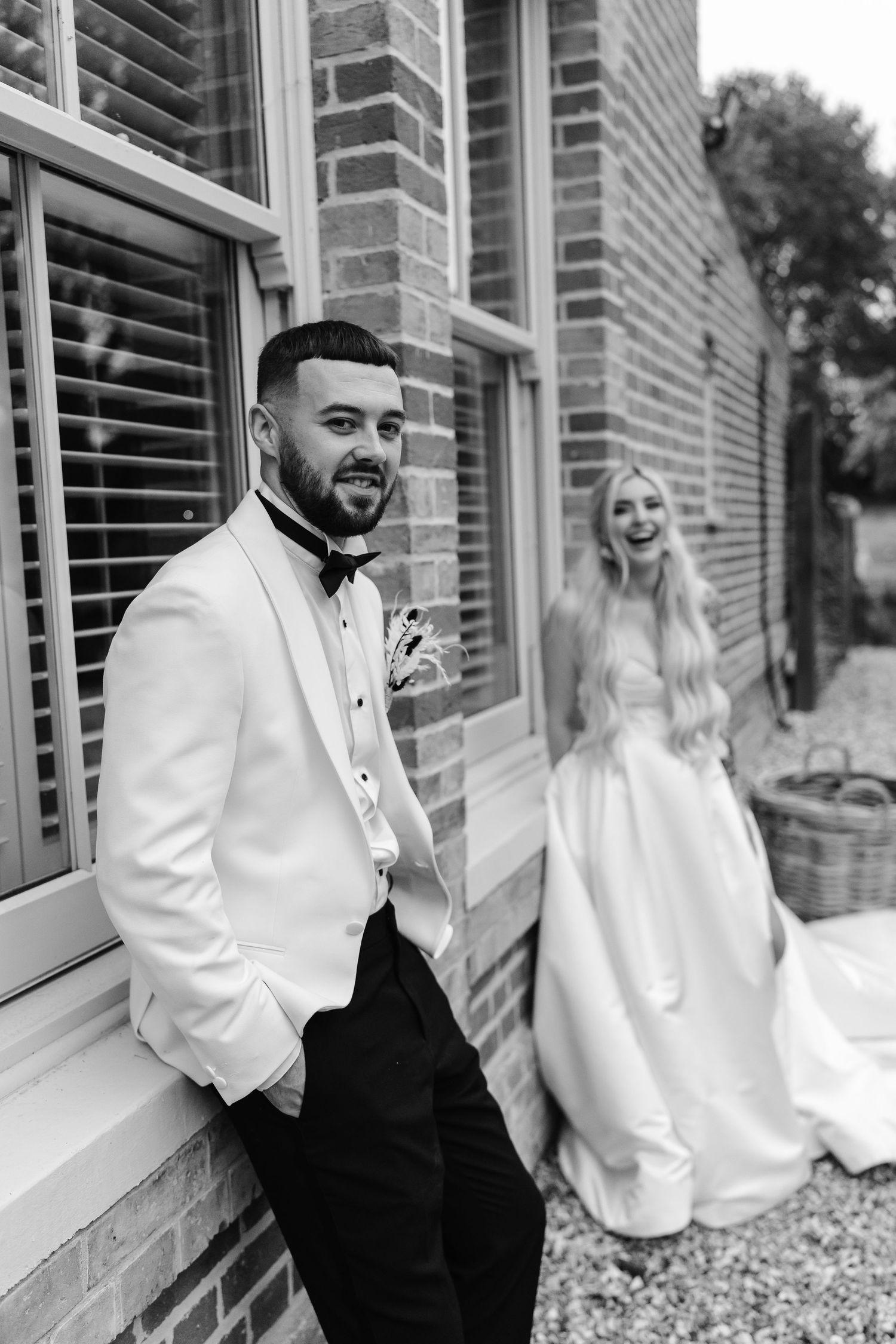 Black and white portrait of groom in white tuxedo leaning against brick wall with smiling bride in background.