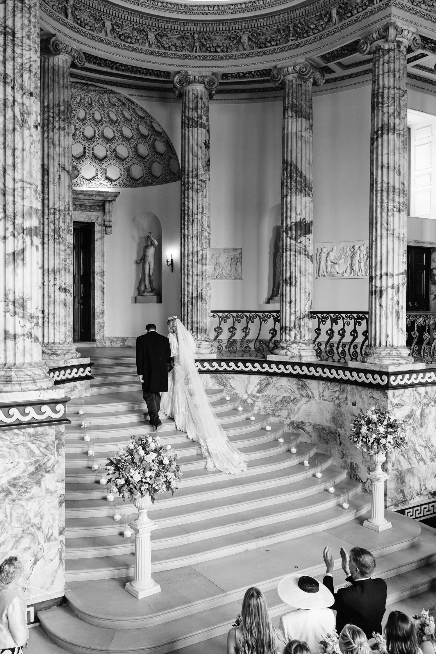 Black and white photo of bride and groom ascending ornate marble staircase in grand neoclassical wedding venue.