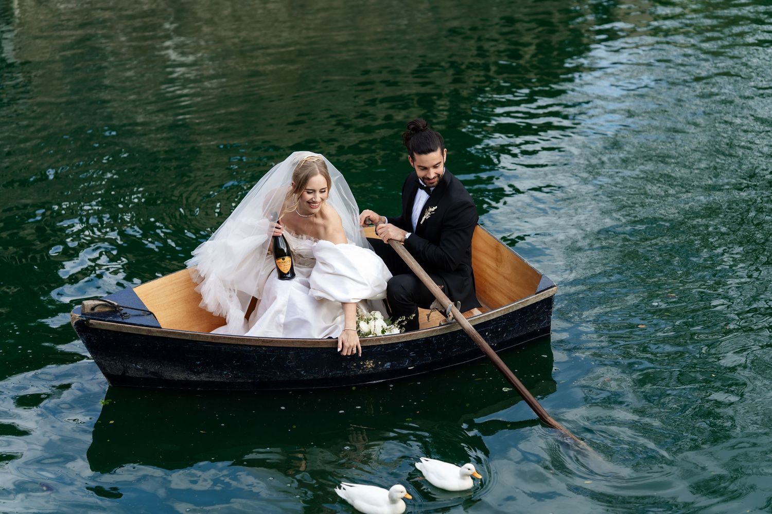 Bride and groom rowing a wooden boat on a green lake, celebrating with champagne on their wedding day.