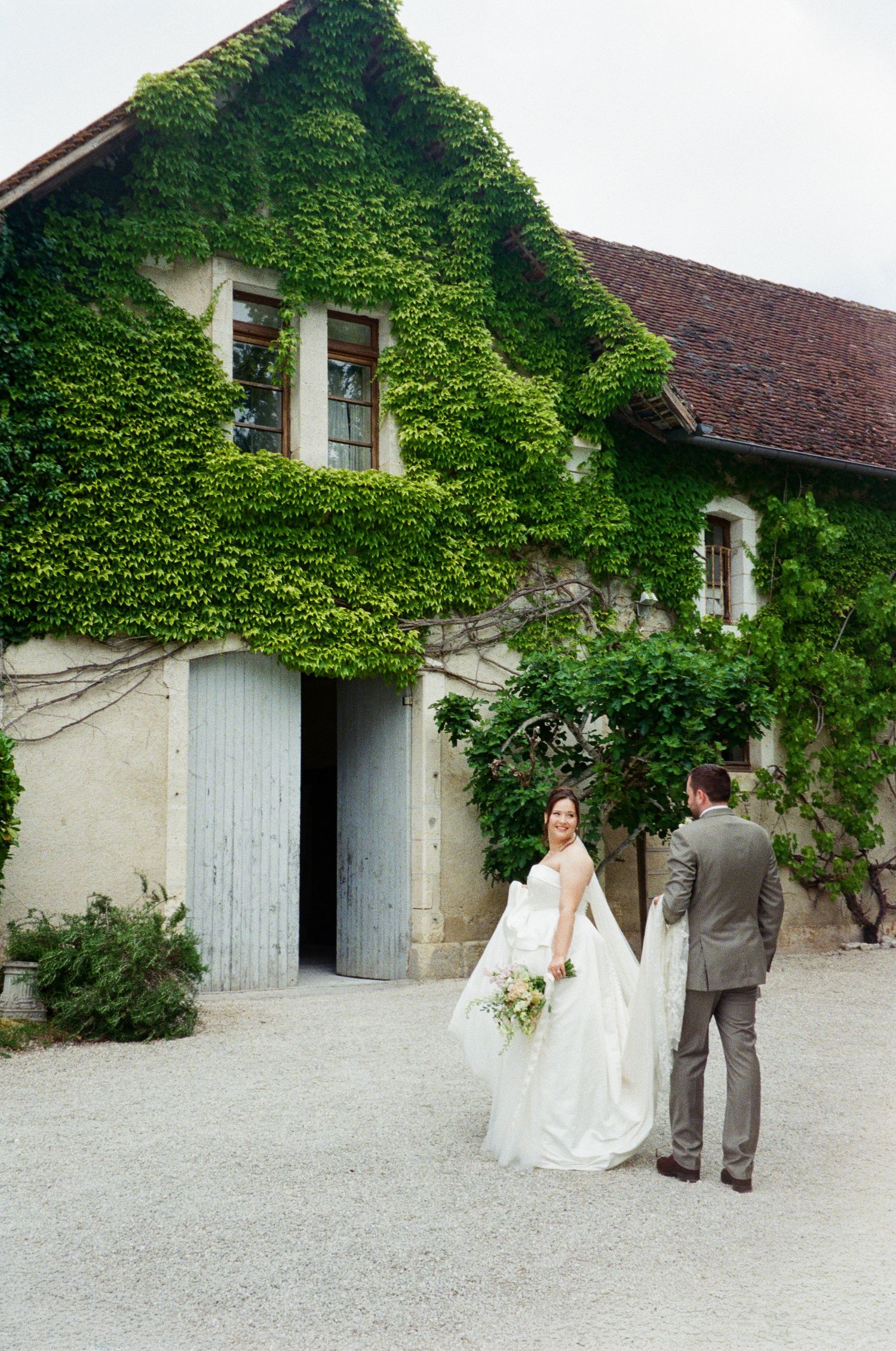 A bride and groom walk hand in hand toward an ivy-covered stone farmhouse on their wedding day.