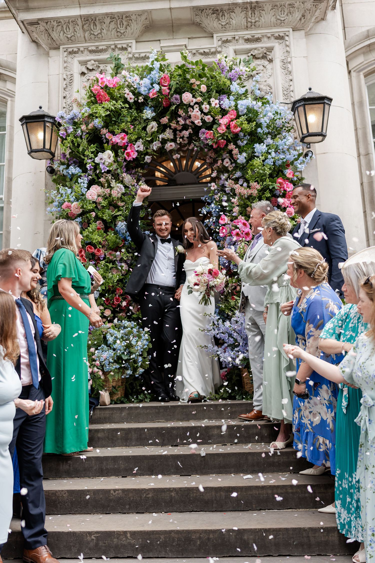 Newlyweds exit grand floral doorway as guests shower them with white confetti on steps.