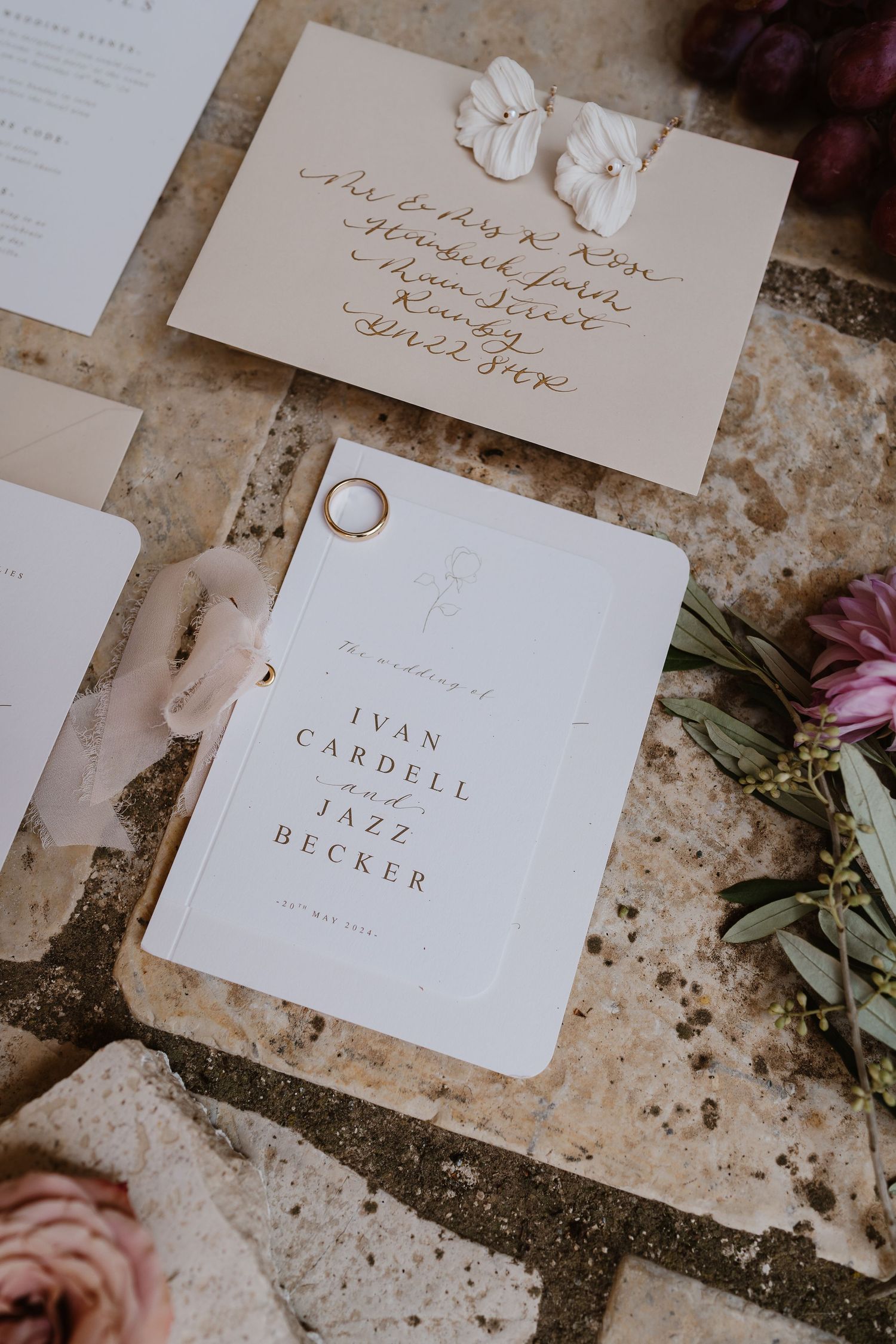 Elegant wedding stationery flat lay featuring gold calligraphy envelope, program, ring, and fresh florals.