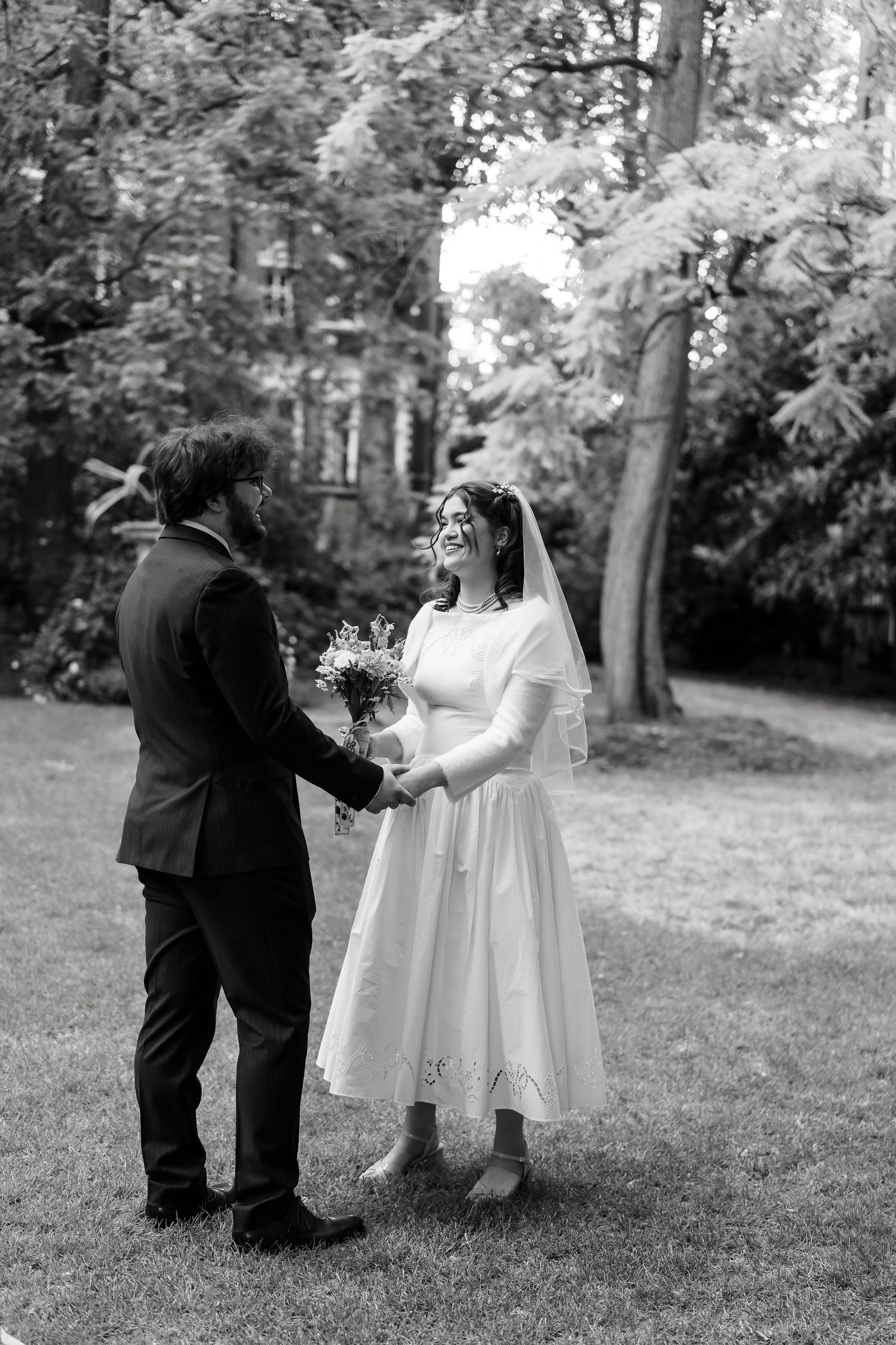 Black and white photo of bride and groom holding hands in lush garden, bride wearing midi veil dress holding bouquet.